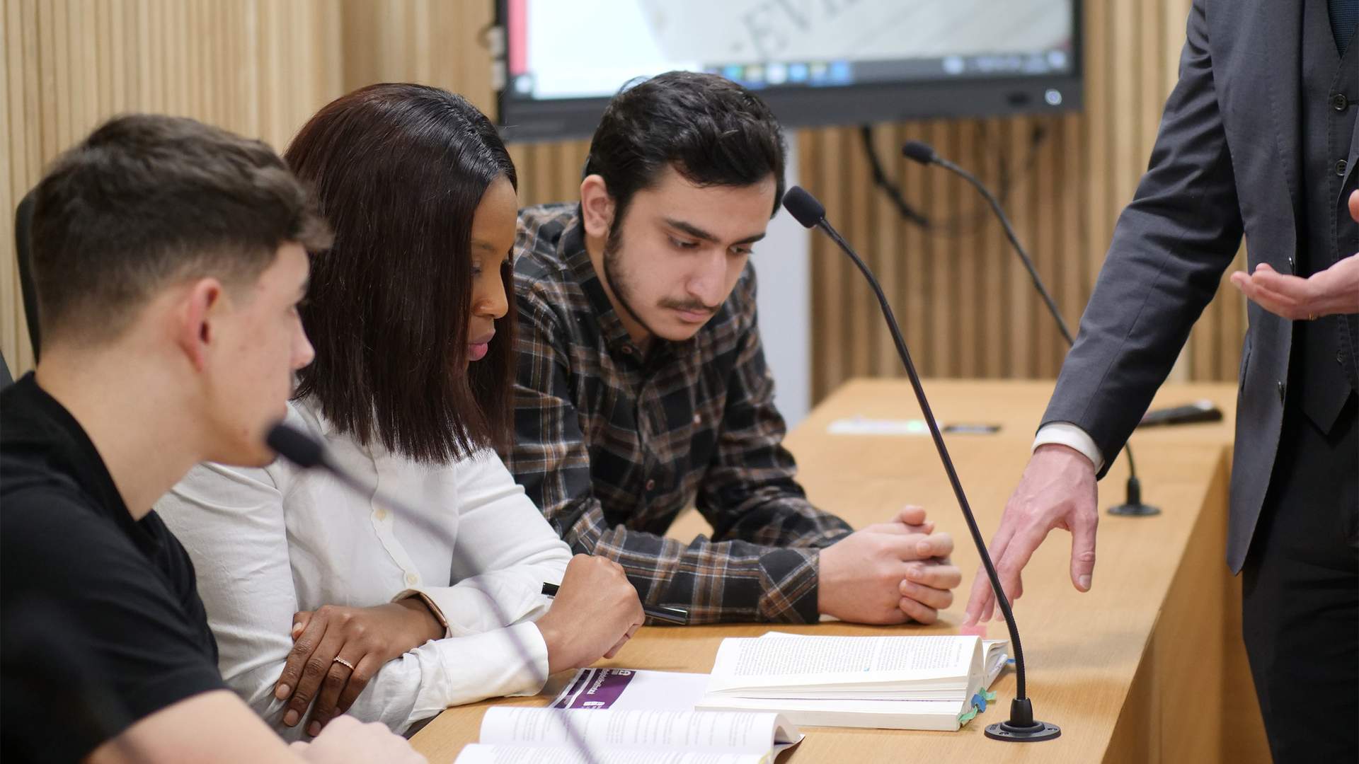 Four people are gathered around a table with open books and microphones, engaging in a discussion. A person in a suit stands nearby, gesturing with their hand. A screen in the background displays a blurred document with the word EVIDENCE.