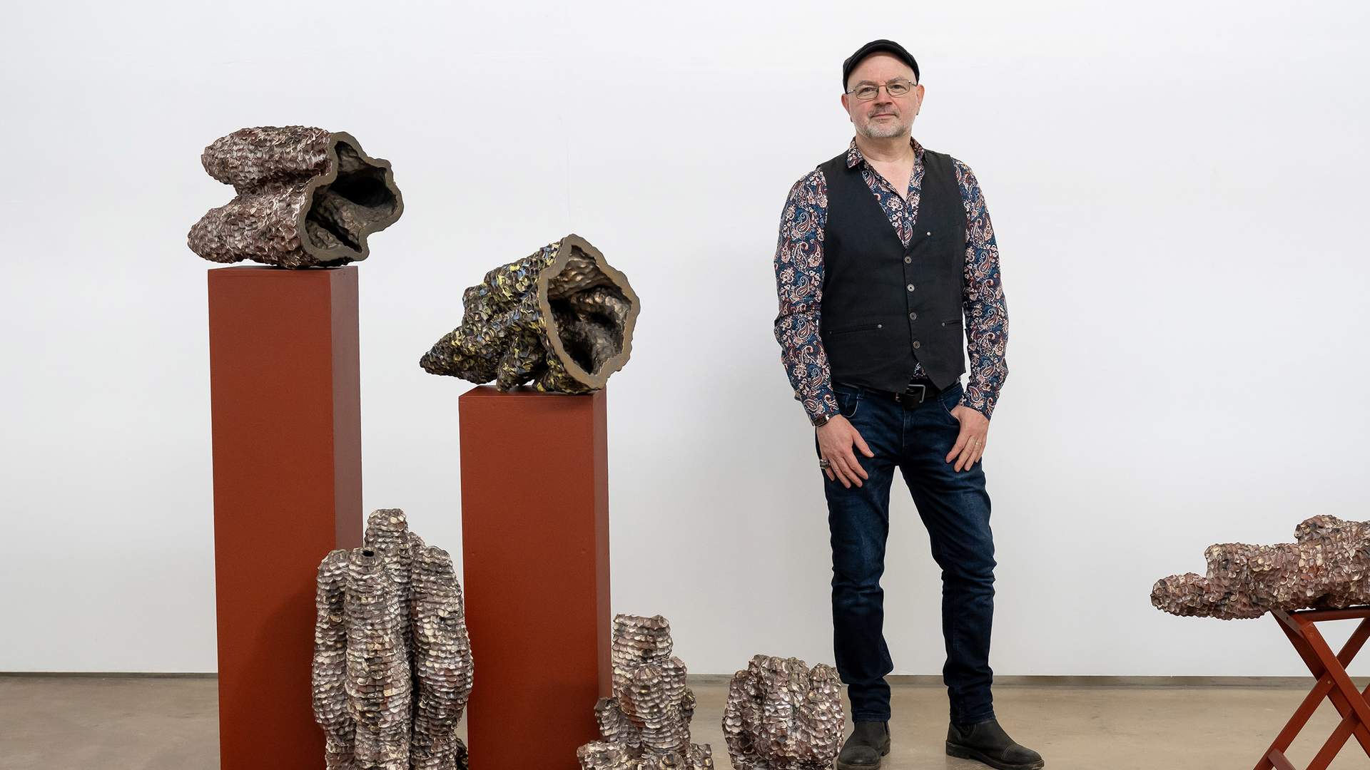 A man stands in a gallery next to various abstract sculptures resembling organic forms. The sculptures are displayed on reddish-brown pedestals against a plain white wall. He is wearing a patterned shirt, vest, jeans, and a cap.
