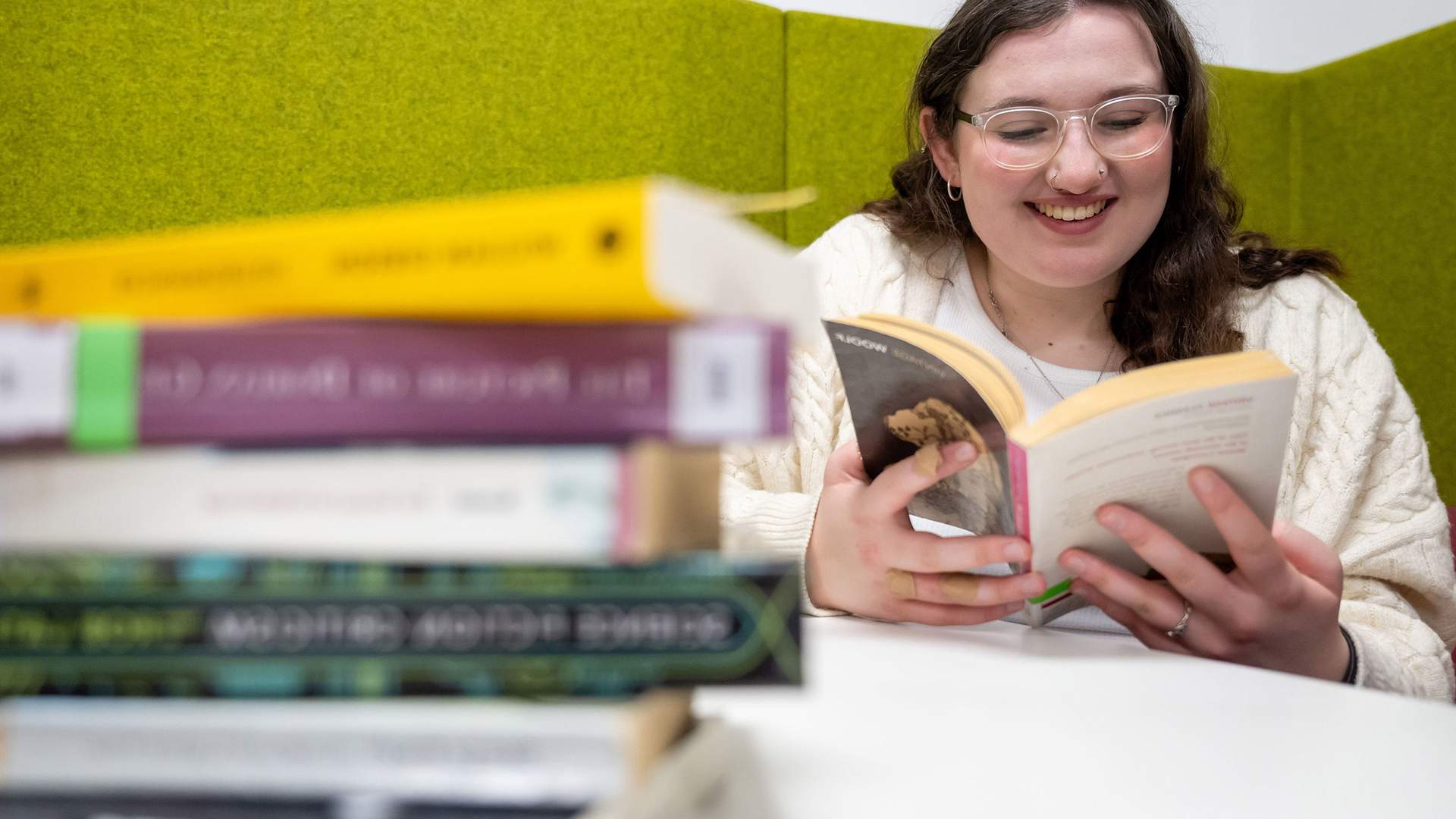 A person with glasses and long hair is smiling while reading a book at a white table. A stack of various books is in the foreground. The background features green padded walls.