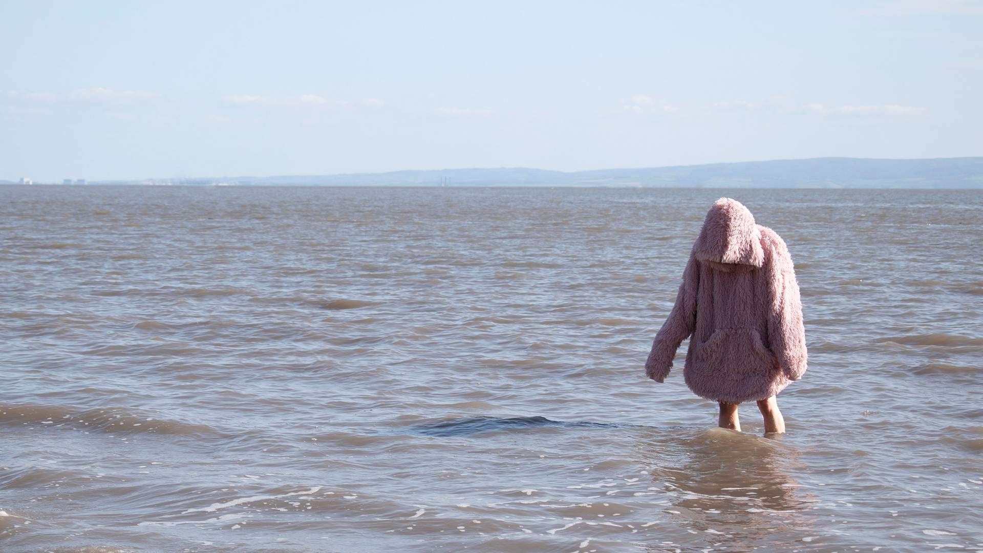A person in a fluffy pink coat wades into the calm sea under a clear sky, with distant hills on the horizon.