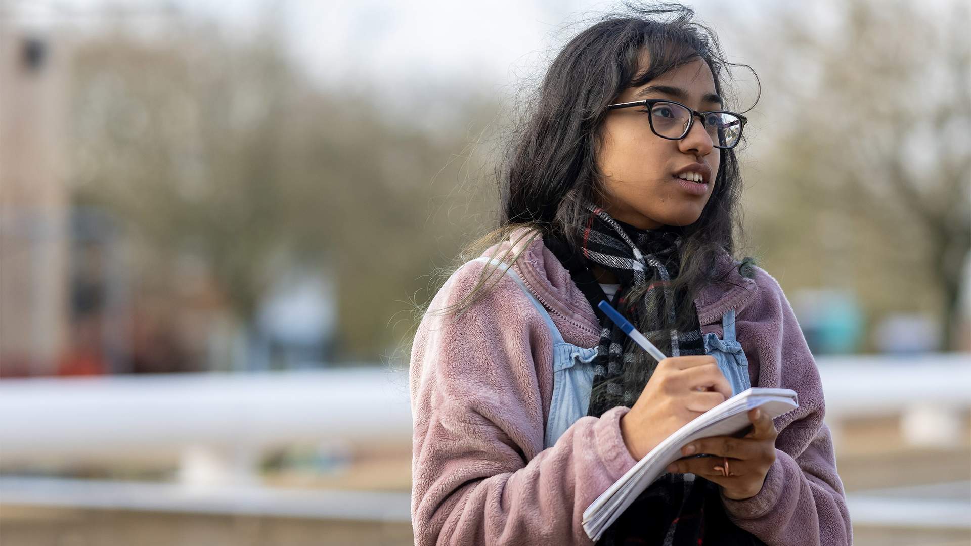 A person with long hair and glasses is outdoors, wearing a pink jacket and holding a notepad and pen. They seem to be taking notes or sketching. The background is blurred with trees and a structure visible.