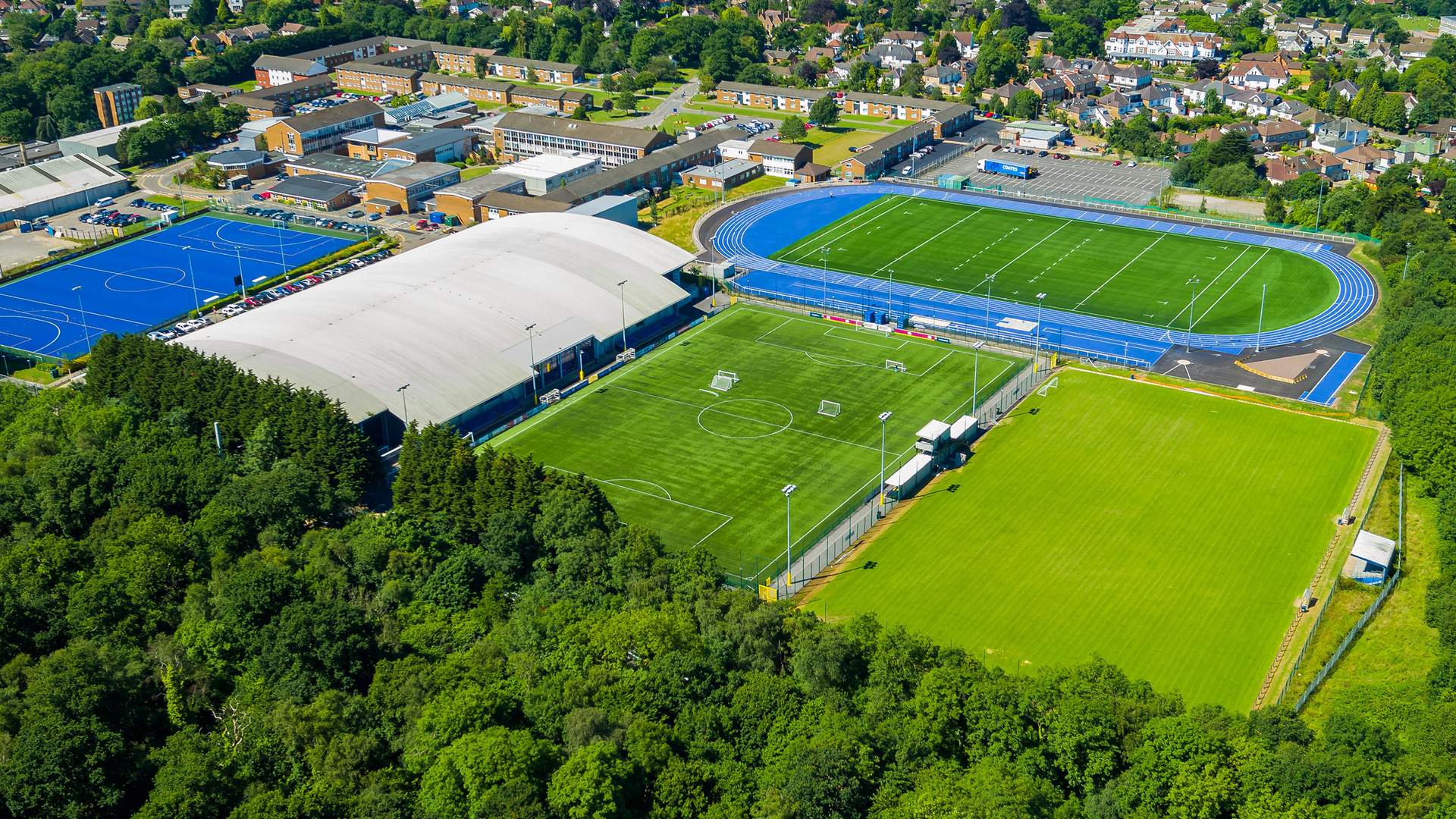 Aerial view of a sports complex with a blue athletics track surrounding a green football field. Adjacent is an indoor arena and another outdoor field, surrounded by lush trees and urban buildings in the background.