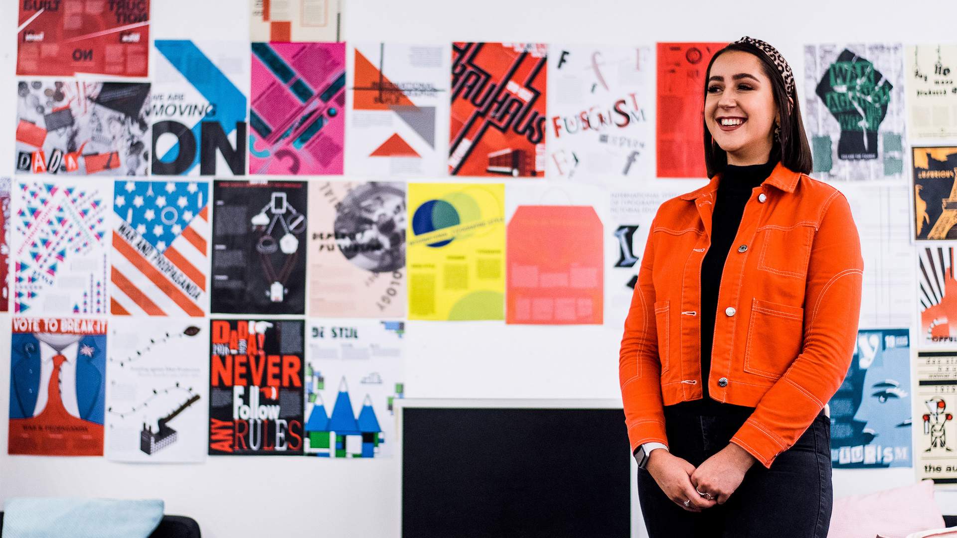 A person in an orange jacket stands smiling in front of a wall filled with colorful, abstract art posters. Theres a monitor and a couch in the foreground.