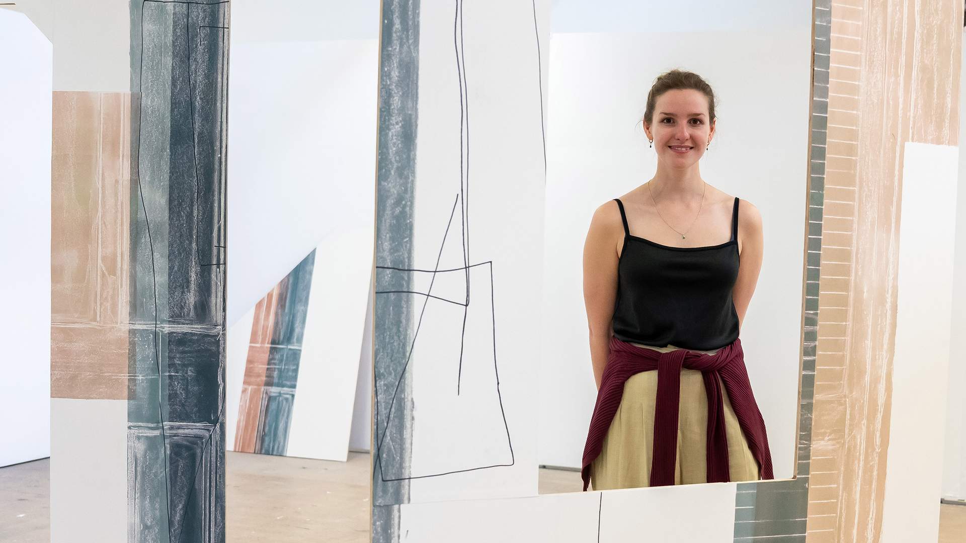 A woman stands smiling amidst an abstract art installation with geometric designs on large panels. She is wearing a black top and has a sweater tied around her waist. The setting appears to be an art gallery.