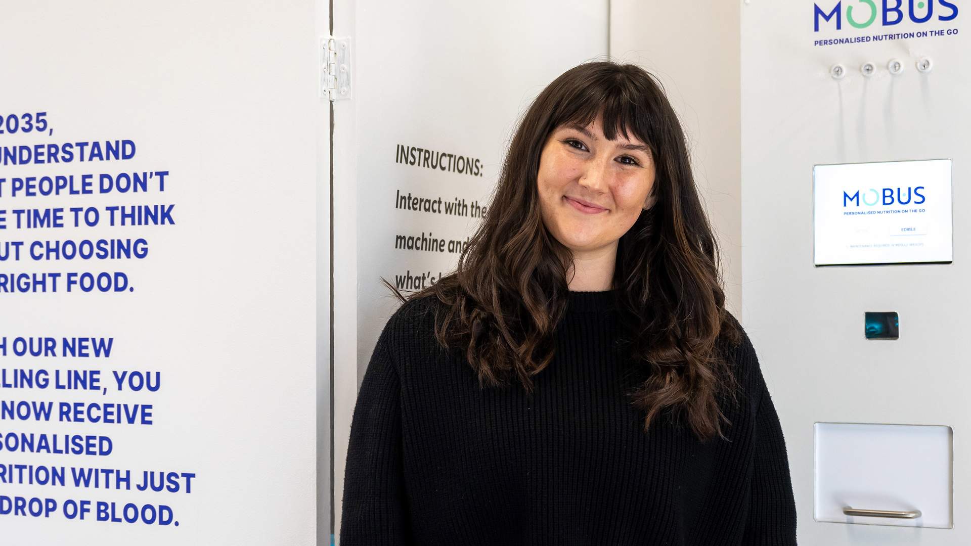 A person with long dark hair is smiling in front of a white vending machine labeled MOBUS. Text on the wall partially reads about understanding nutritional needs by 2035. The person is wearing a black sweater.