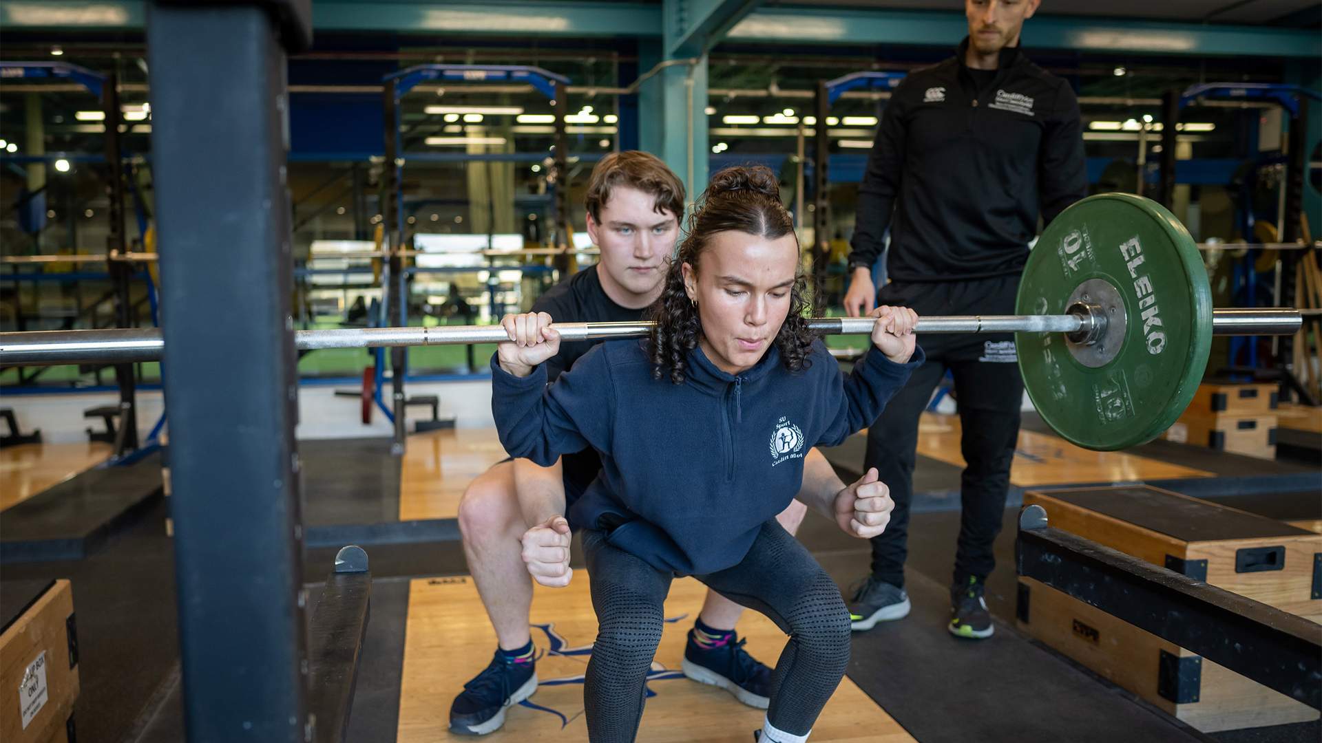 A person is performing a squat with a barbell in a gym. Another person behind them is spotting to assist, while a third person observes the exercise. The gym has weightlifting equipment and wooden platforms.