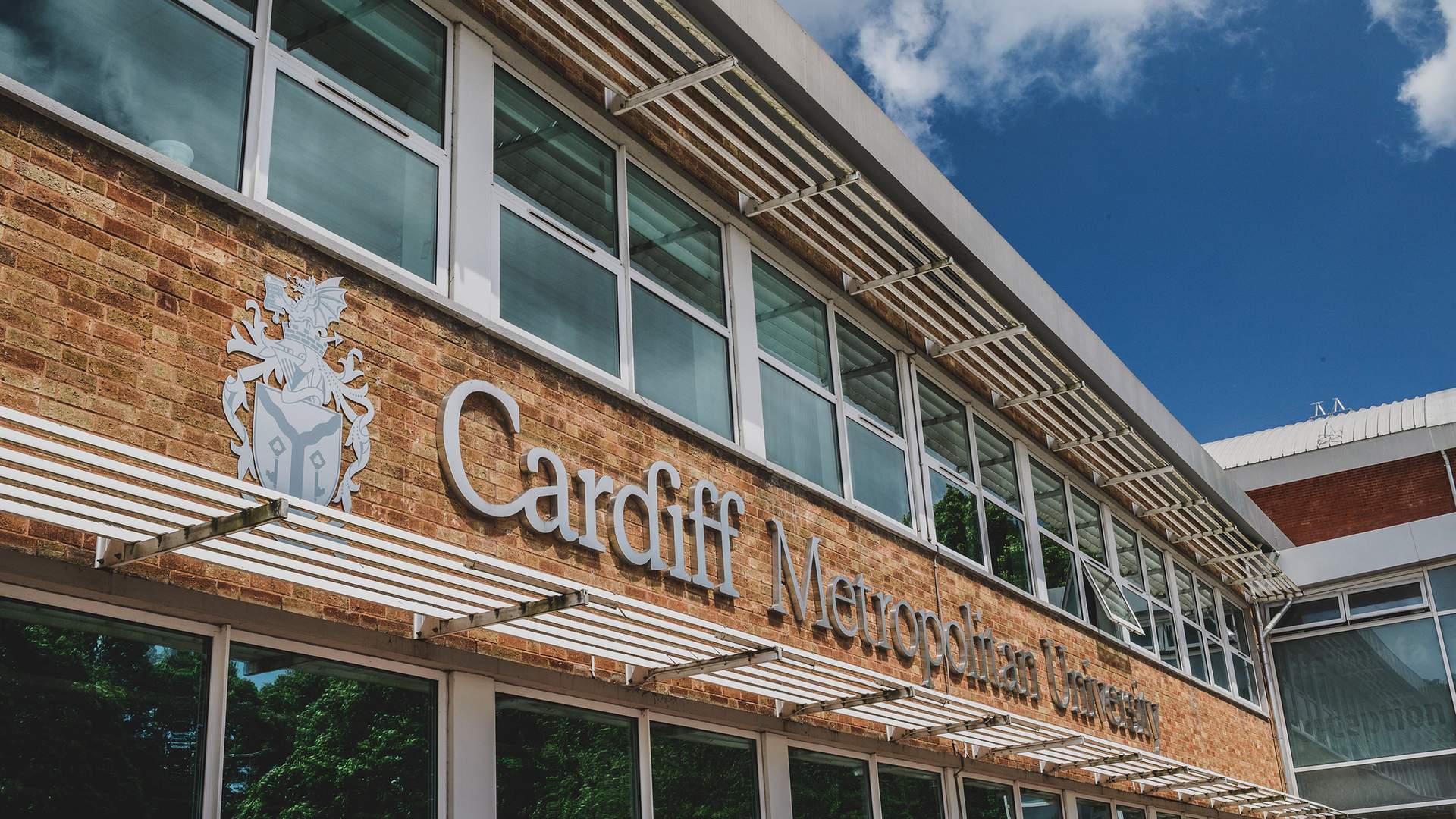 The image shows the exterior of Cardiff Metropolitan University. The building has a modern design with large windows and the universitys name prominently displayed in white lettering on a brick facade under a blue sky.