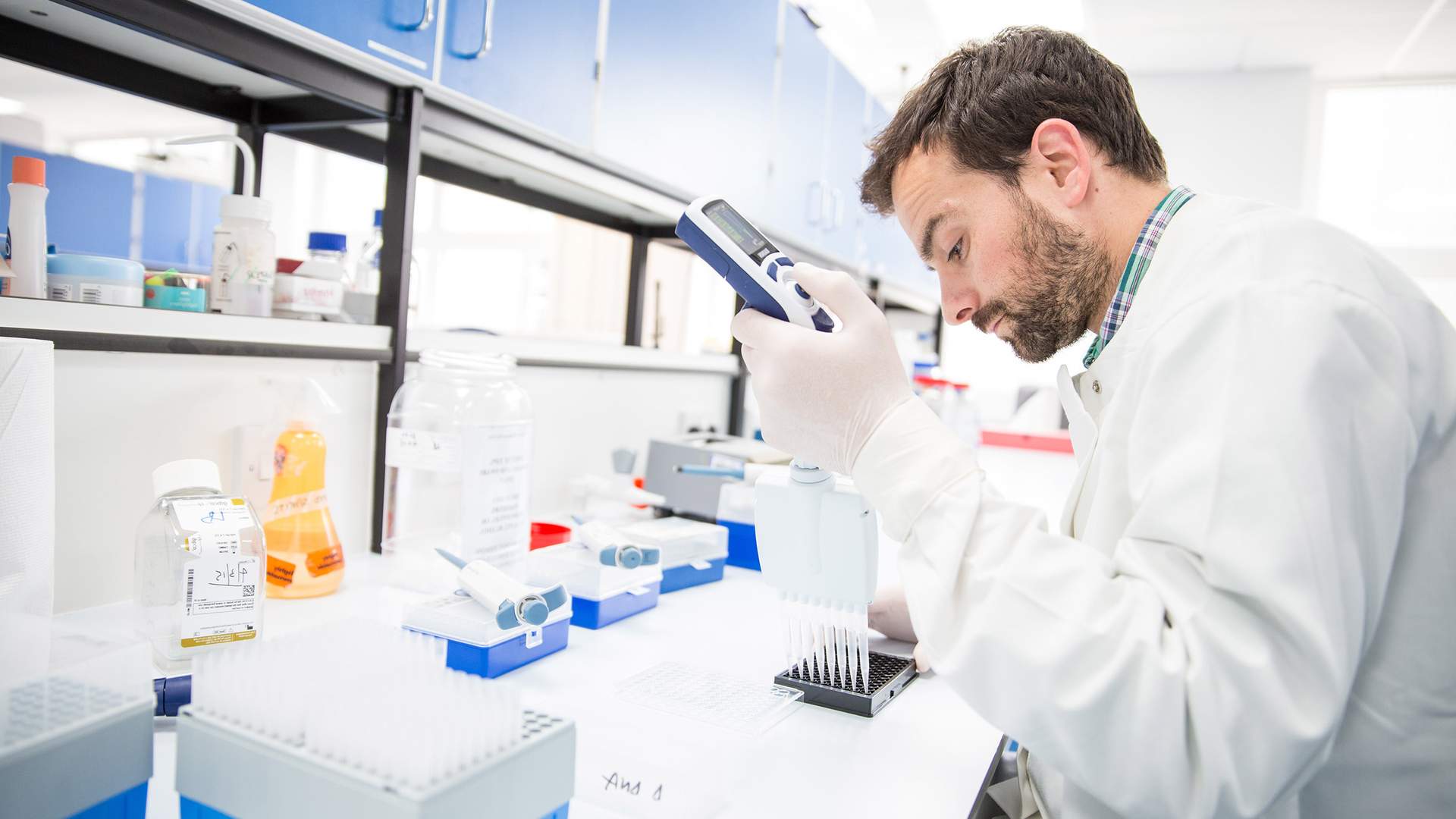 A scientist in a lab coat uses a pipette to transfer liquid into a tray, surrounded by lab equipment and bottles.