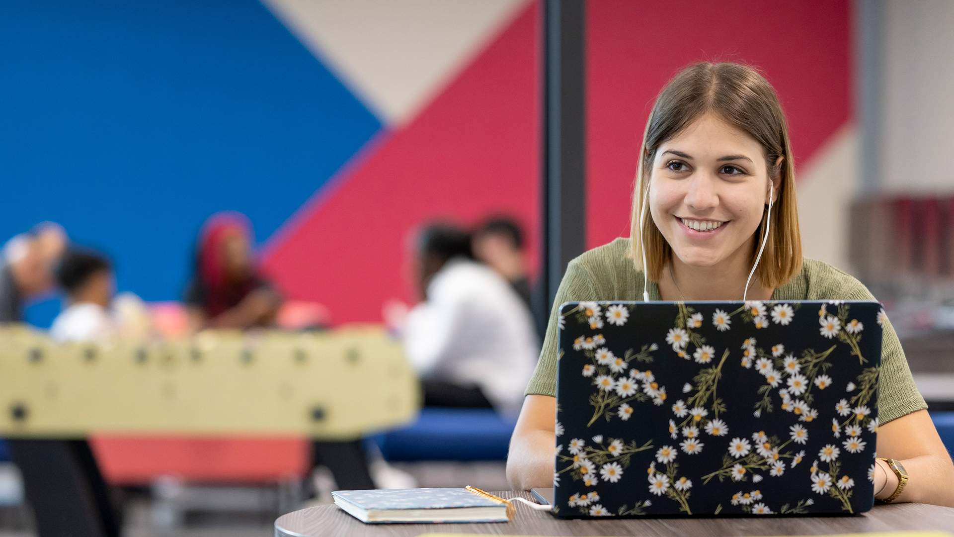 A woman smiling while using a laptop in a colorful, casual workspace. She is wearing earbuds and sitting at a round table. In the background, several people are gathered, and a foosball table is visible.
