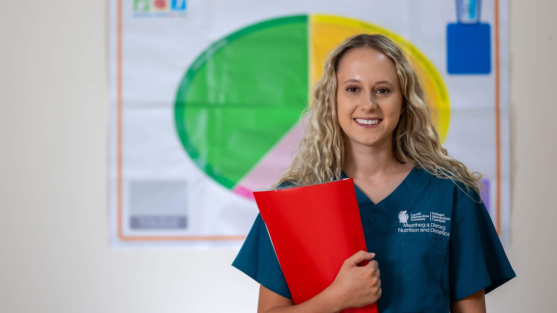A woman in a teal uniform smiles while holding a red folder. She stands in front of a colorful chart, likely related to nutrition or dietetics. Her long blonde hair is loose, and she appears confident and professional.