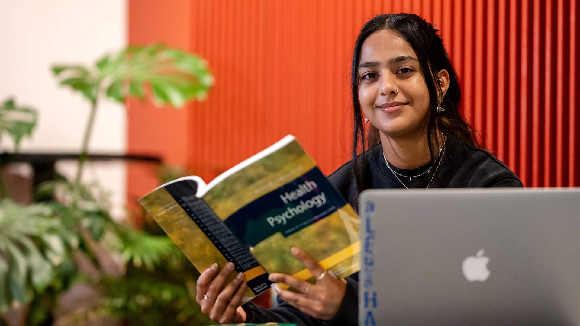 A person sits at a table with an open book titled Health Psychology and a laptop in front of them. They are smiling and positioned against a backdrop with red and orange vertical lines. Green plants are visible in the background.