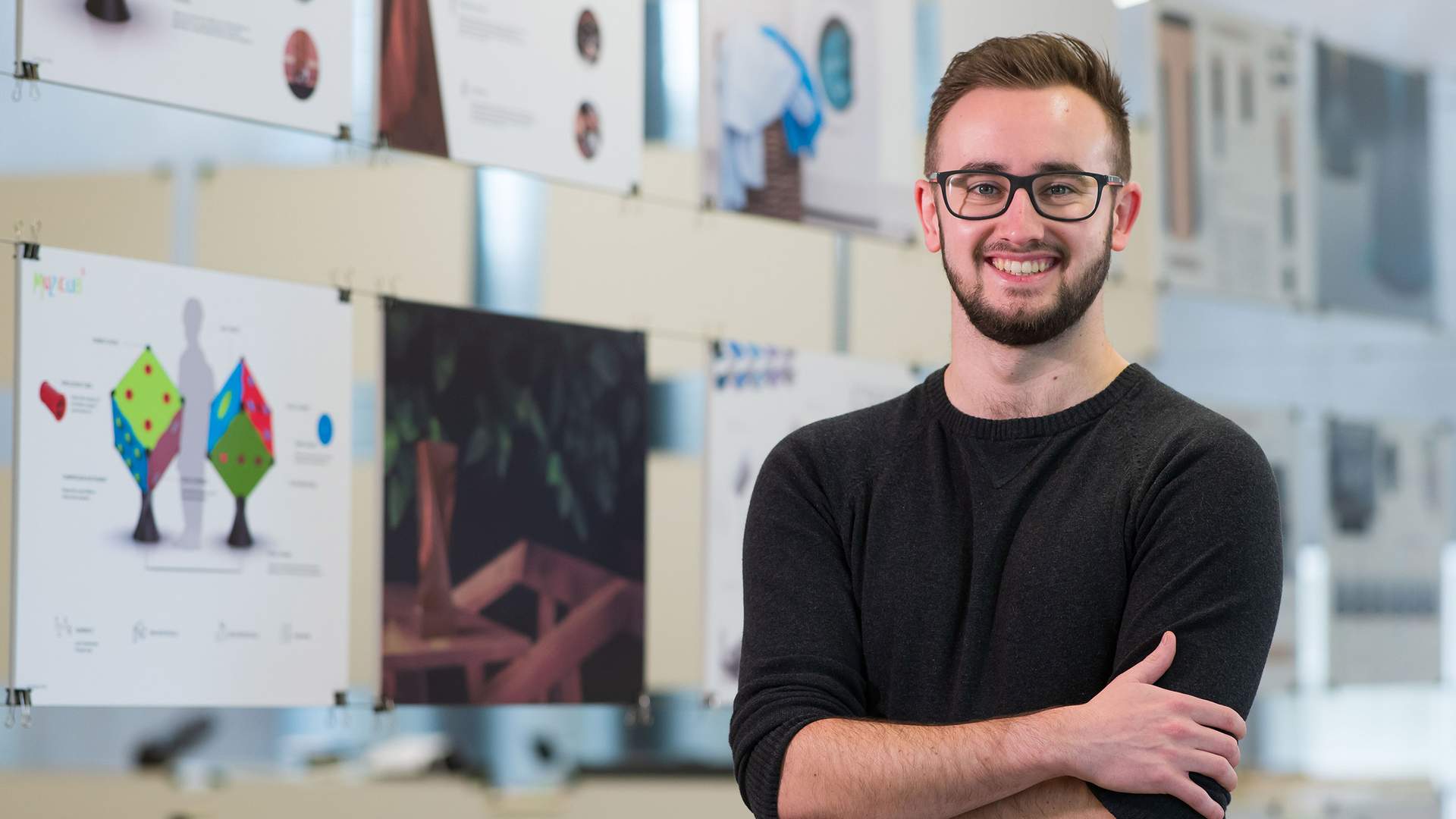 A man wearing glasses and a black sweater stands smiling with his arms crossed in front of display boards featuring various design projects and abstract images. The background is a well-lit room with natural light.