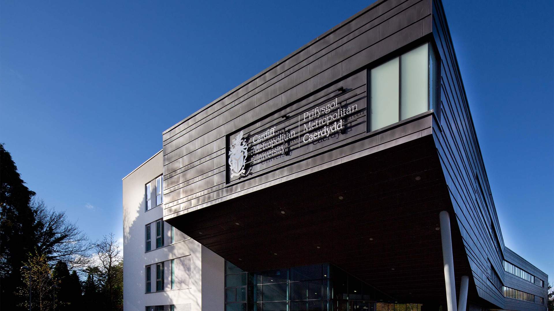 Modern building with angular architecture, featuring a large sign that reads Prifysgol Metropolitan Caerdydd. The structure has a mix of dark panels and white walls, with glass windows. Trees and a blue sky are visible in the background.