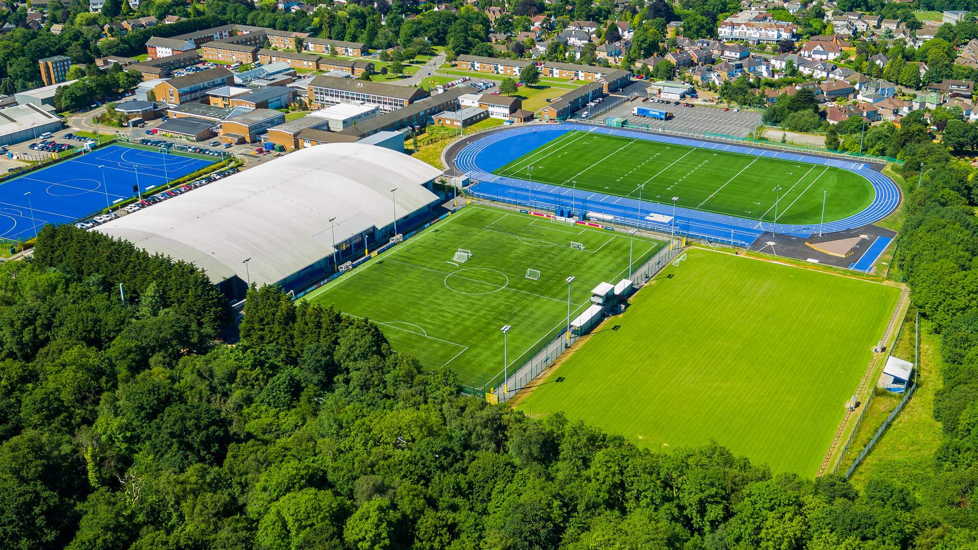 Aerial view of an athletic complex featuring a green soccer field, blue running track, and domed sports facility. Surrounding the complex are trees and residential buildings in the background.