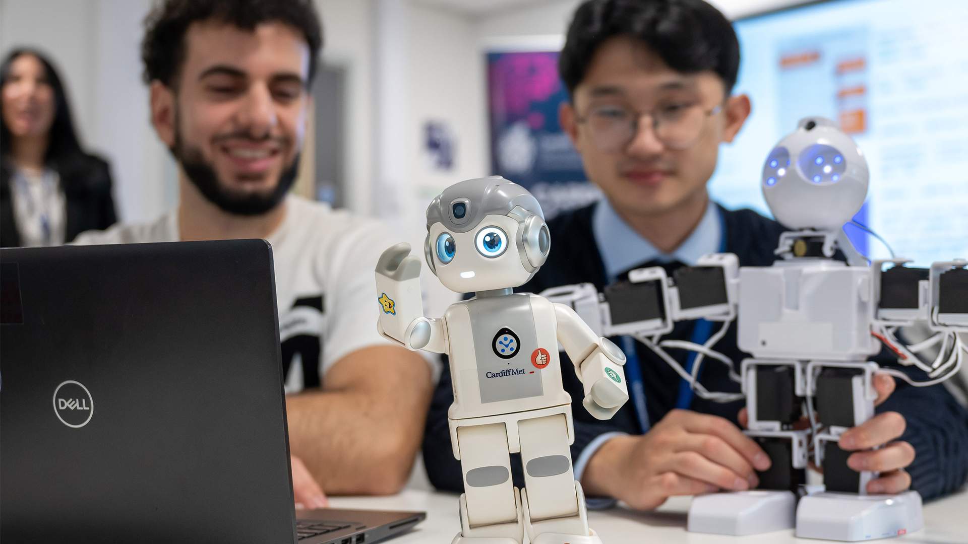 Two men interacting with robots in a tech setting. One is using a laptop, while two humanoid robots are on the table. The foreground robot is small and white with a delighted expression. The background has monitors and a blurred person.