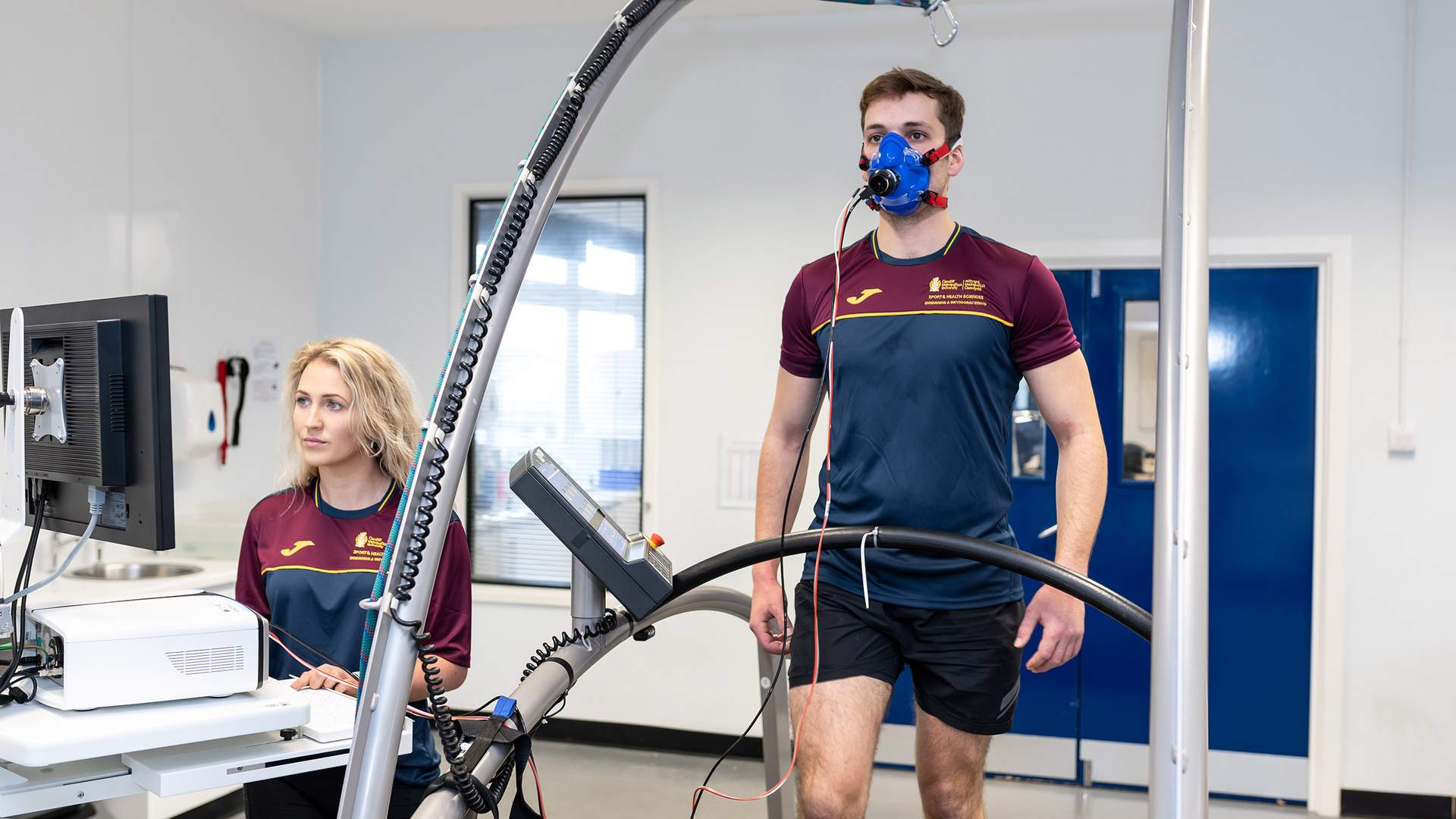 A man is walking on a treadmill wearing a blue mask for a fitness test. A woman sits nearby, operating a computer connected to the equipment. Both are in a lab setting wearing maroon and navy sports attire.