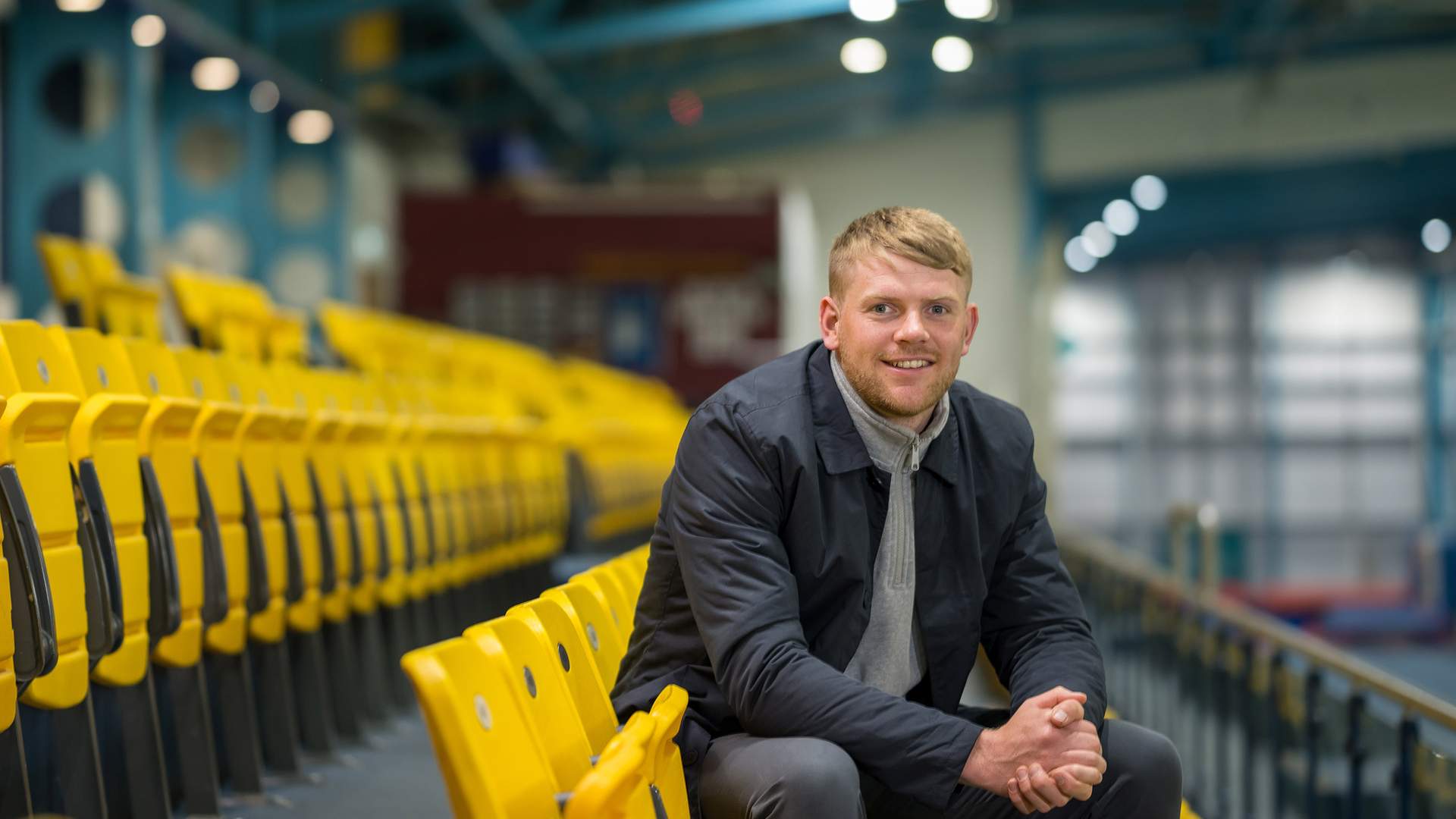 A man sits in an empty sports stadium with yellow seats, wearing a dark jacket and smiling. The background shows a brightly lit gymnasium interior with blue structural beams.