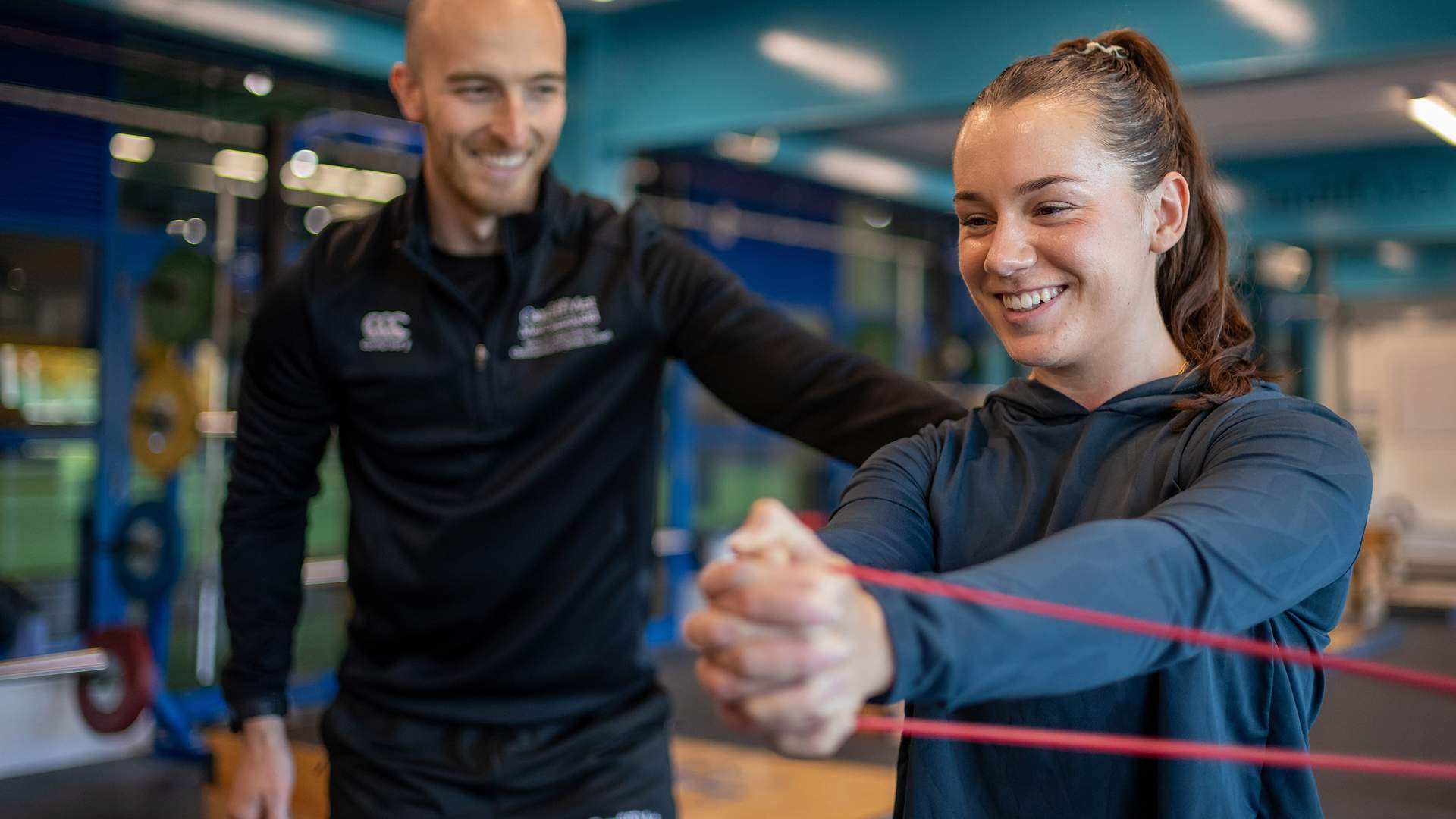 A woman exercises with a red resistance band, held in both hands, as a man in a black tracksuit smiles and assists her. They are in a gym with blue and silver equipment in the background.