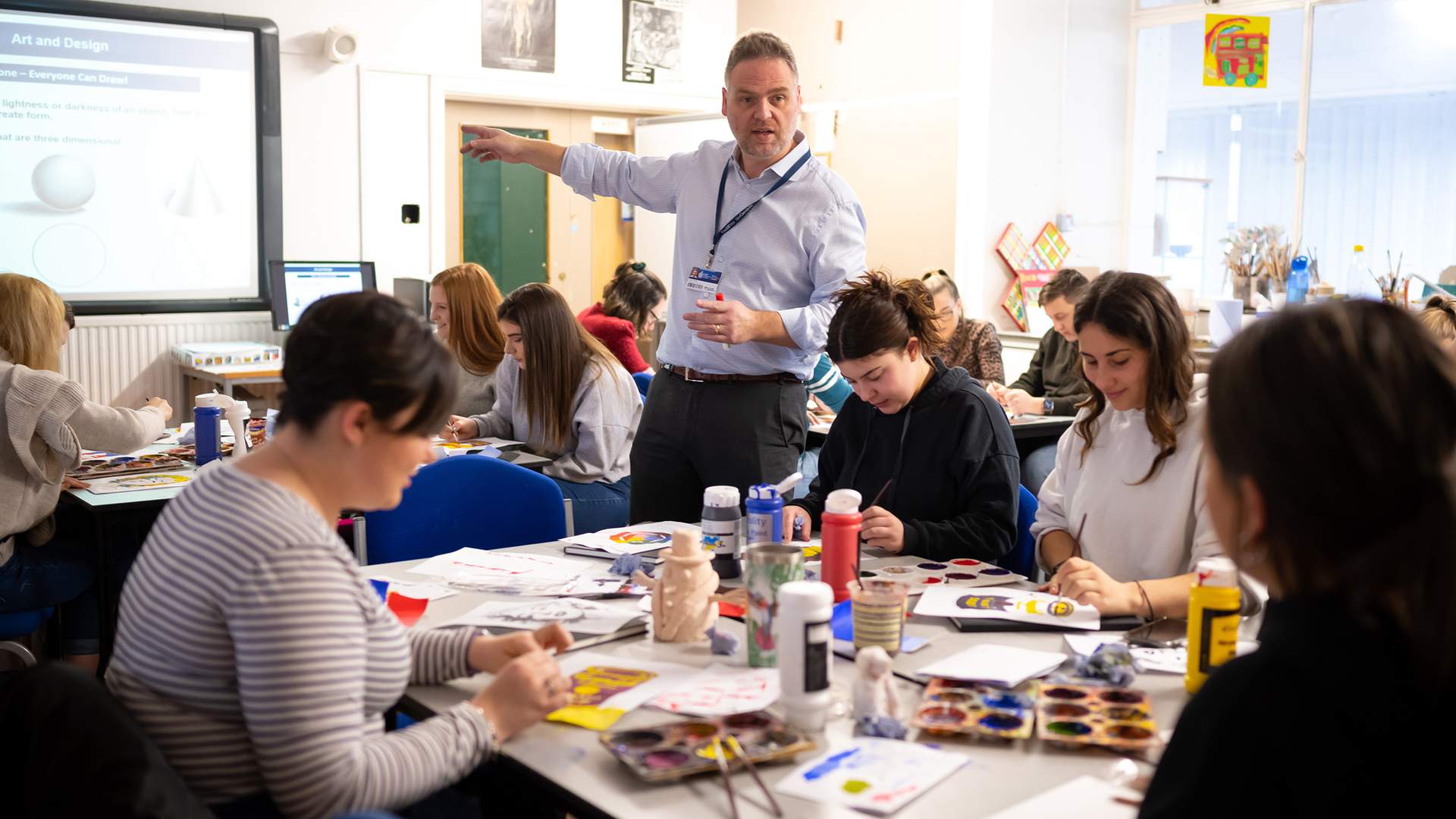 A teacher points at a whiteboard. He is surrounded by pupils at desks with paint and paper.