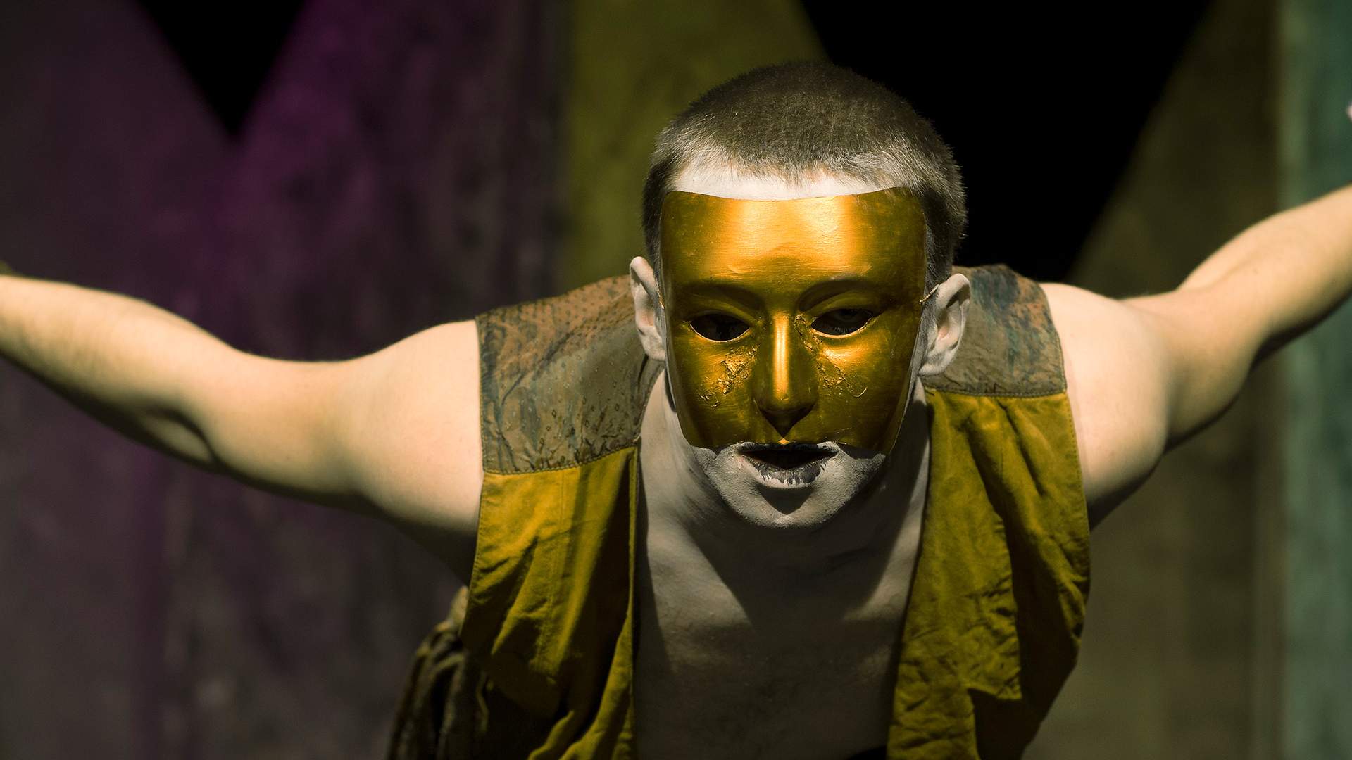 A performer leans forward with arms outstretched. He is wearing a gold mask covering his face.