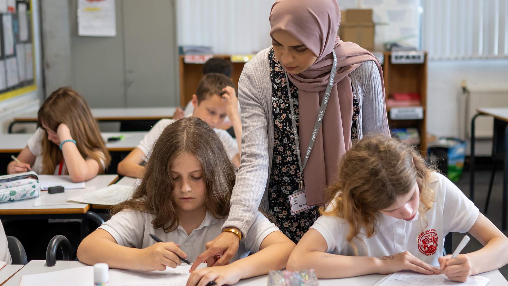A teacher stands over two pupils writing at their desks.