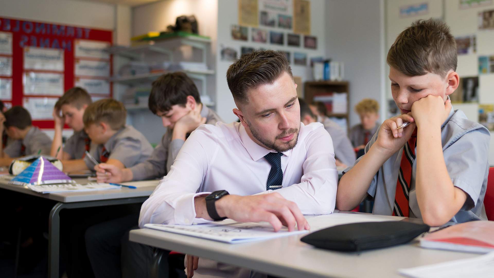 A teacher kneels next to a pupil sitting at a desk. He is pointing to the open page of a book.