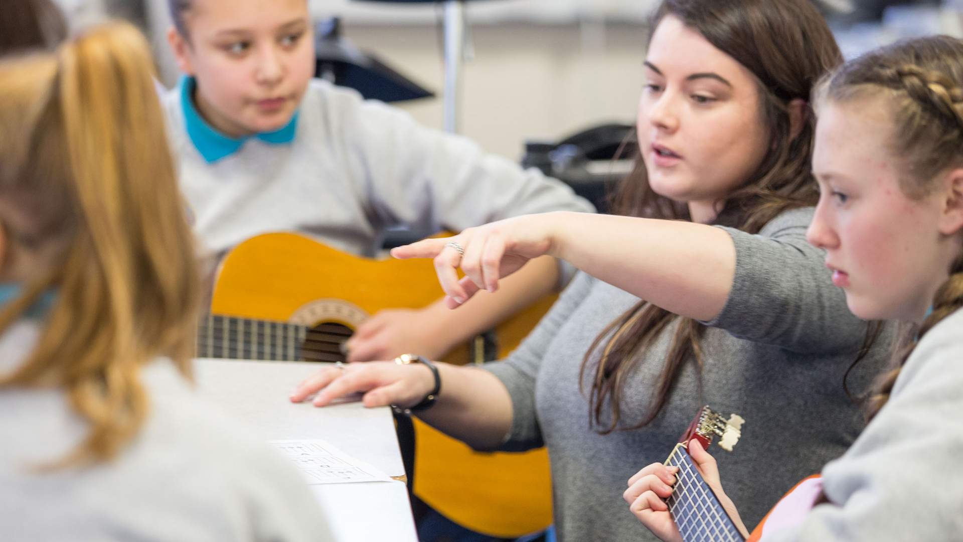 A teacher stands with a group of pupils. Two of the pupils are holding an acoustic guitar.