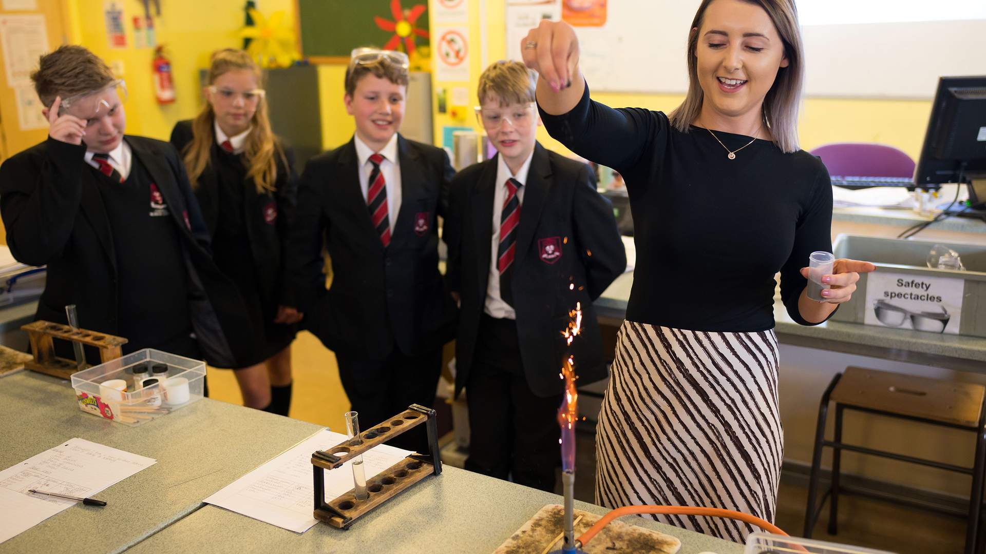 A teacher stands in front of a table. She is dropping powder into the flame of a Bunsen burner.