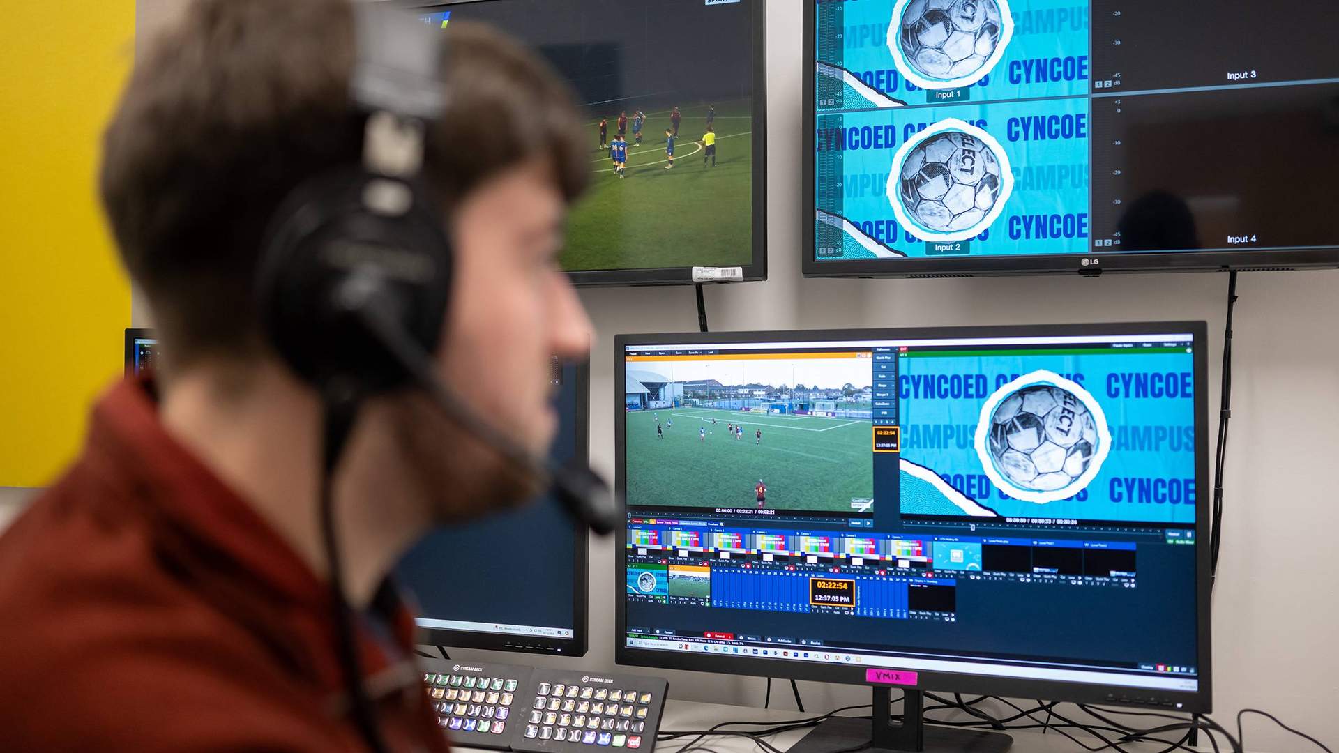 A person in headphones watches a football match on several monitors.