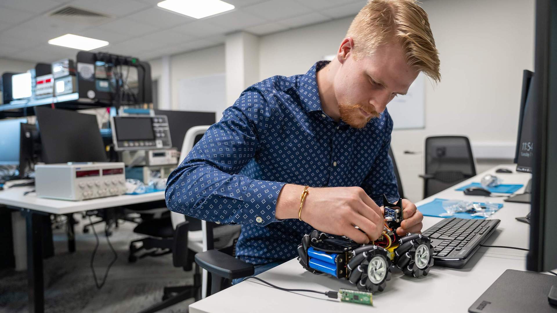 A person assembling a wheeled robot in an electronics lab, surrounded by tools and equipment.
