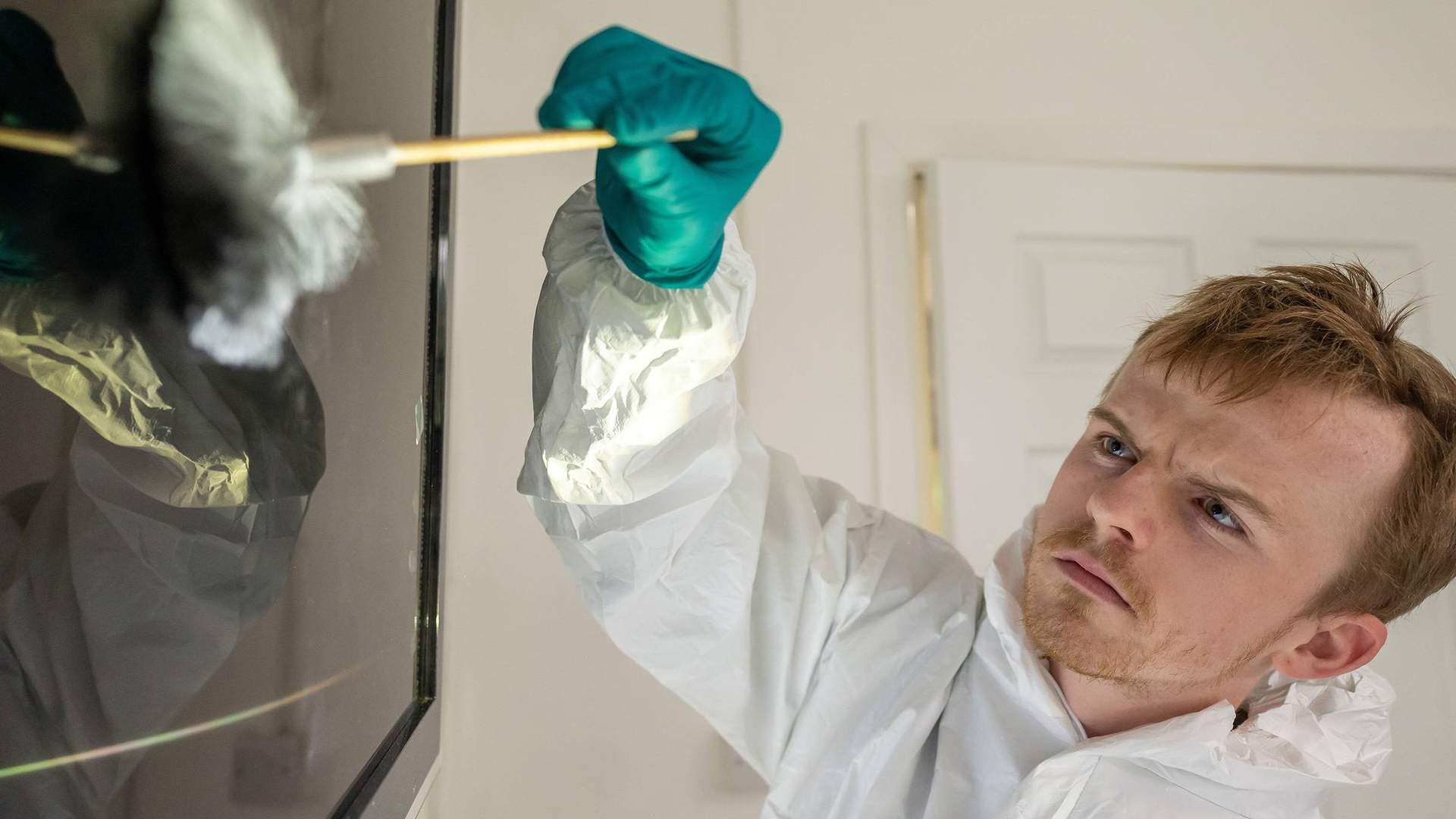 A person wearing a protective suit and teal gloves analyses fingerprints on a reflective surface, marked with green evidence tags.