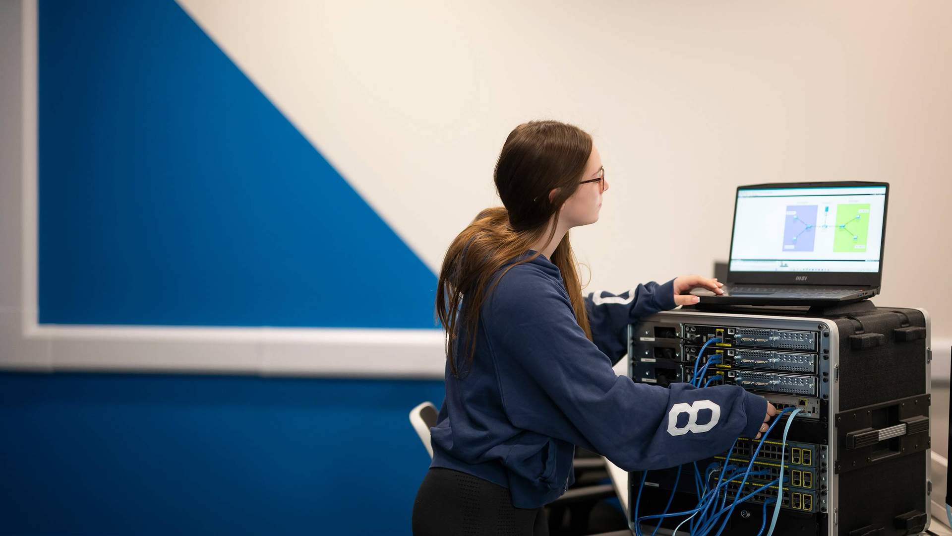 A person plugs cables into computer network equipment while looking at a laptop screen.
