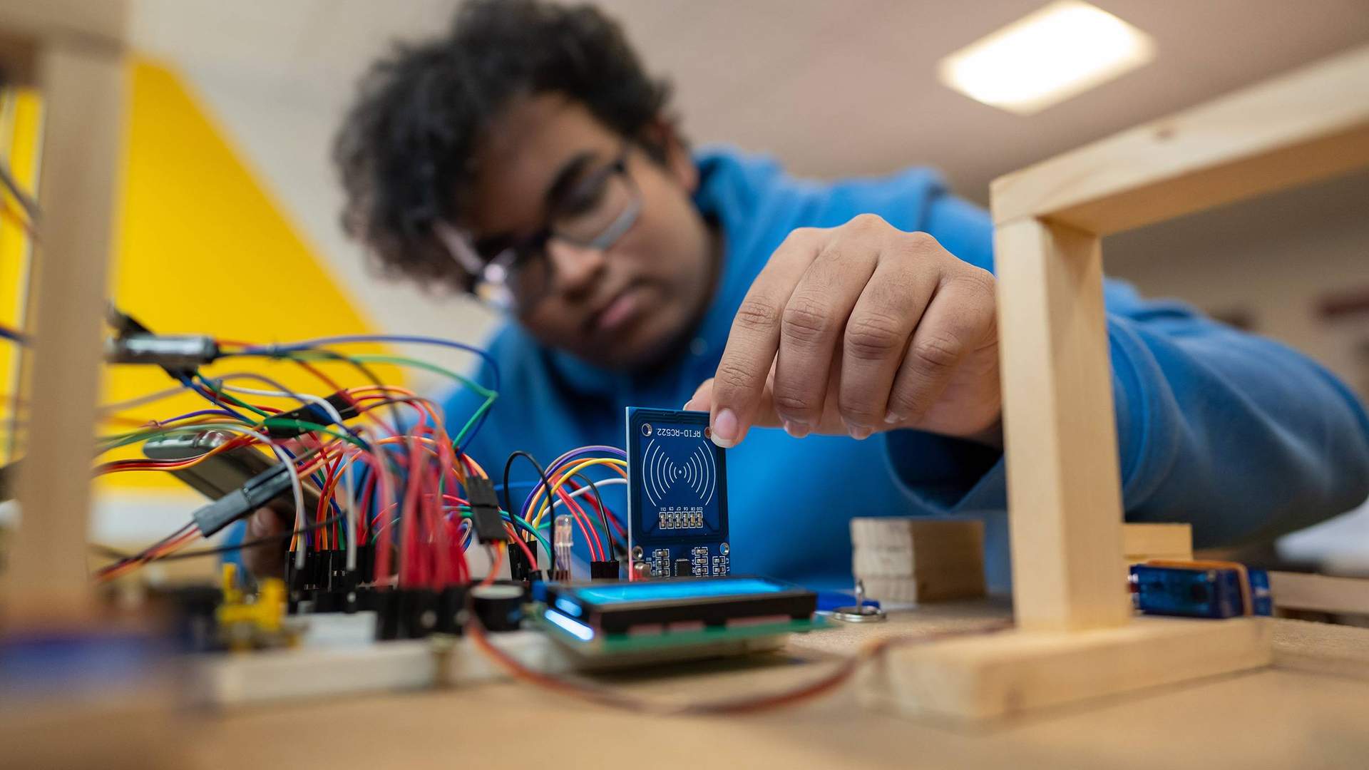 A person in a blue hoodie works with electronic components and circuit boards, placing a small blue board among connecting wires on a table. The background is blurred and the lighting is bright.