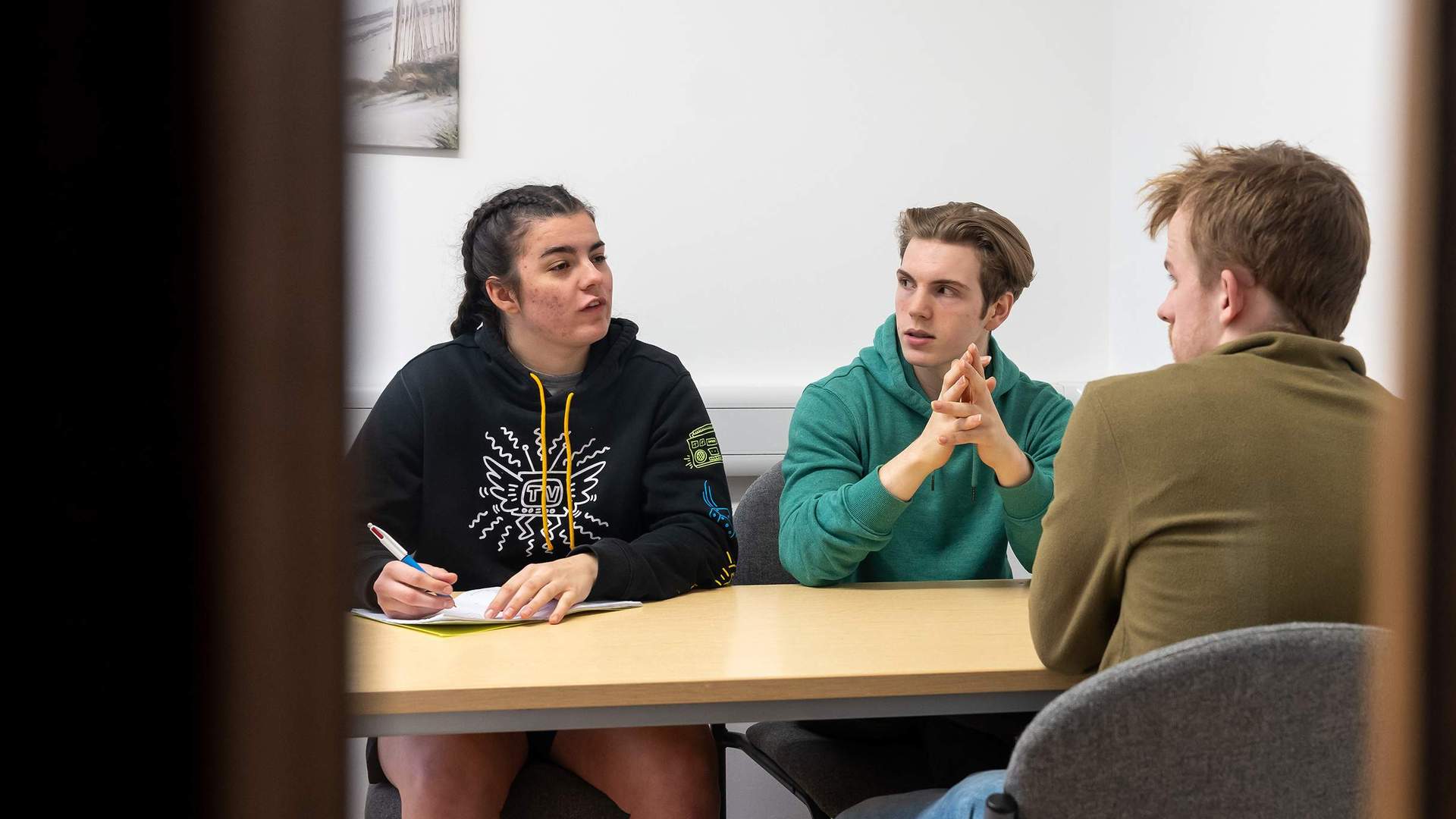 Two people sit at table facing another person, one is writing notes in a notebook.
