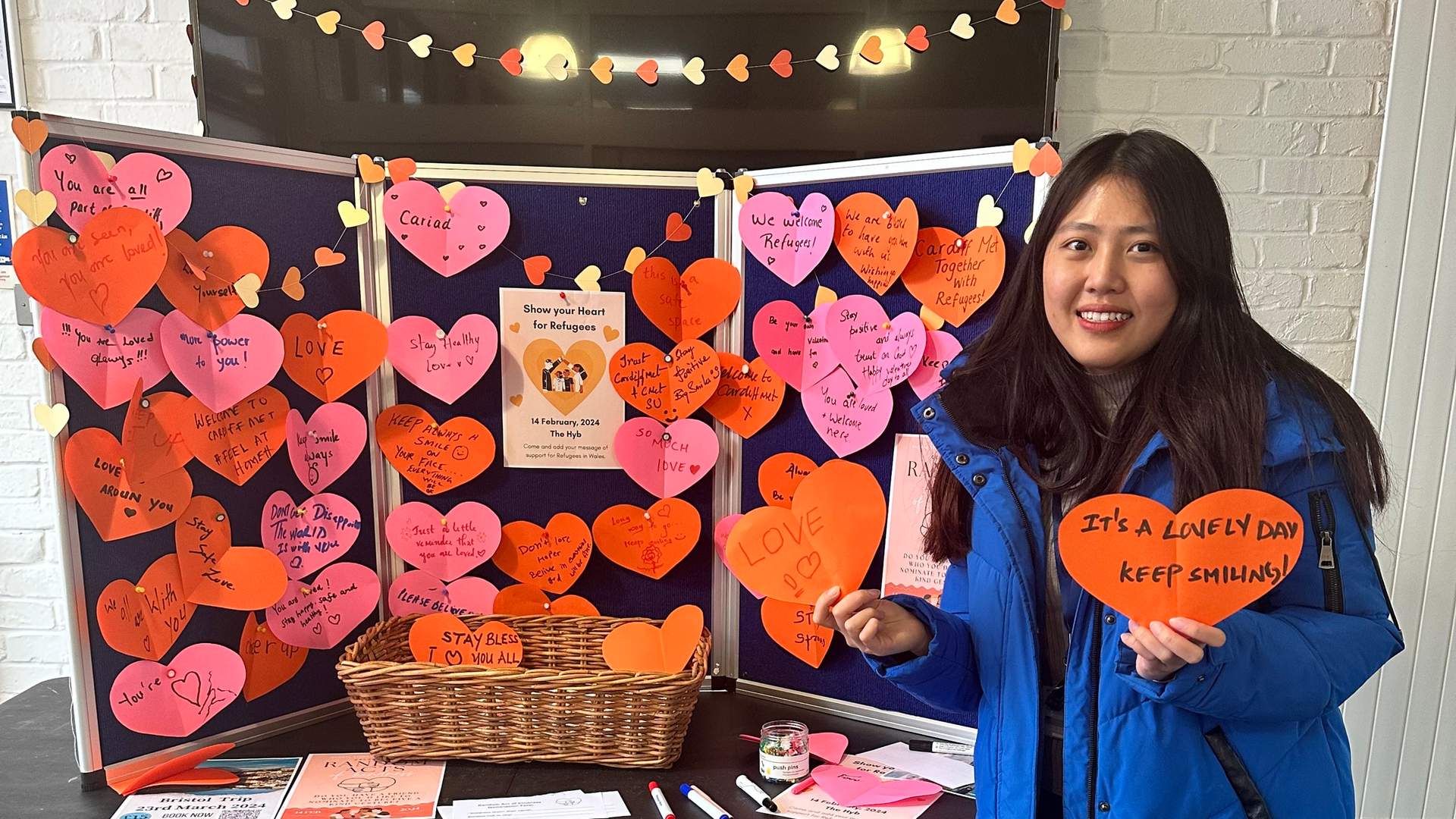 A woman holding a paper heart card stands in front of a display stand filled with paper hearts containing messages of support for refugees