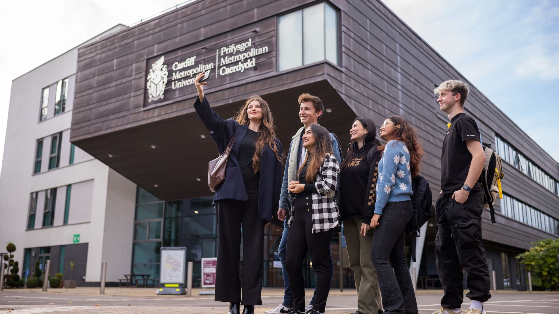 Six young adults stand huddled together outside the Cardiff School of Management as one of them takes a selfie on a mobile phone