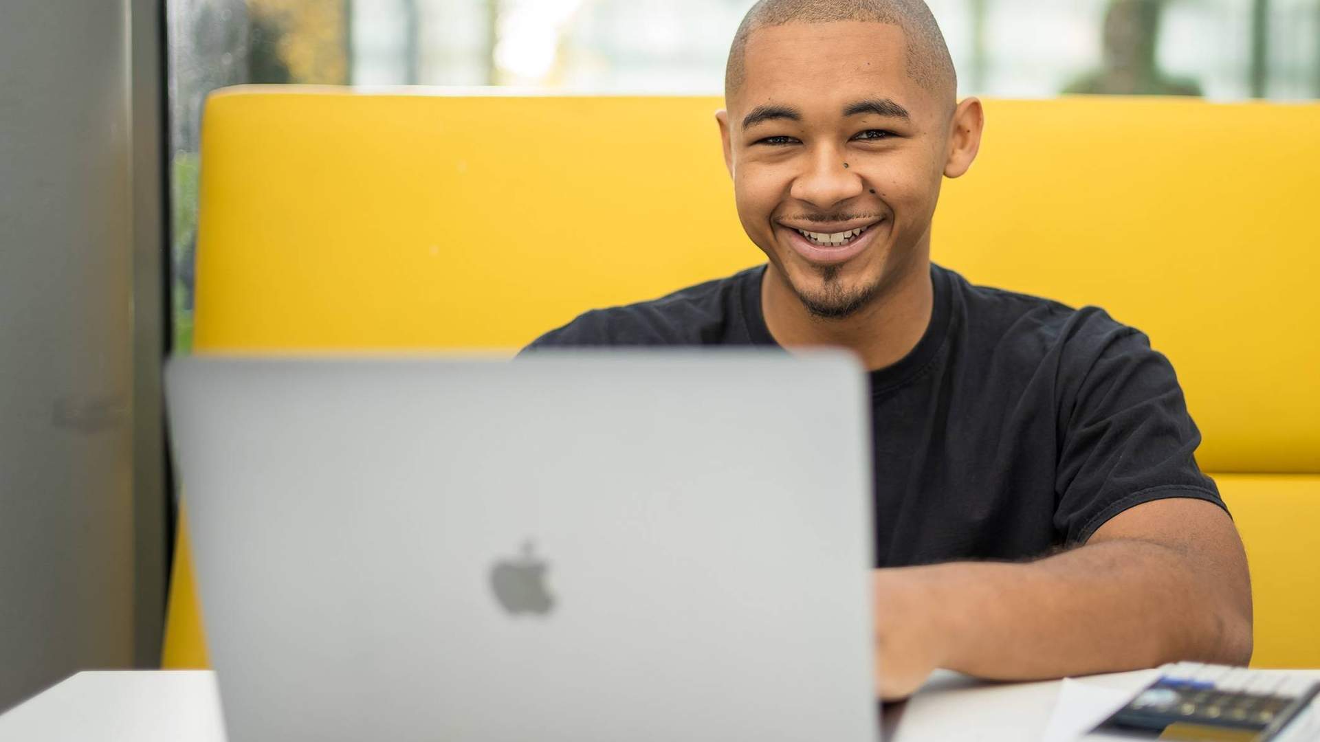 A person is smiling while sitting at a table with a laptop in front of them. They are in a bright, modern setting, with a yellow cushioned seating area behind them.