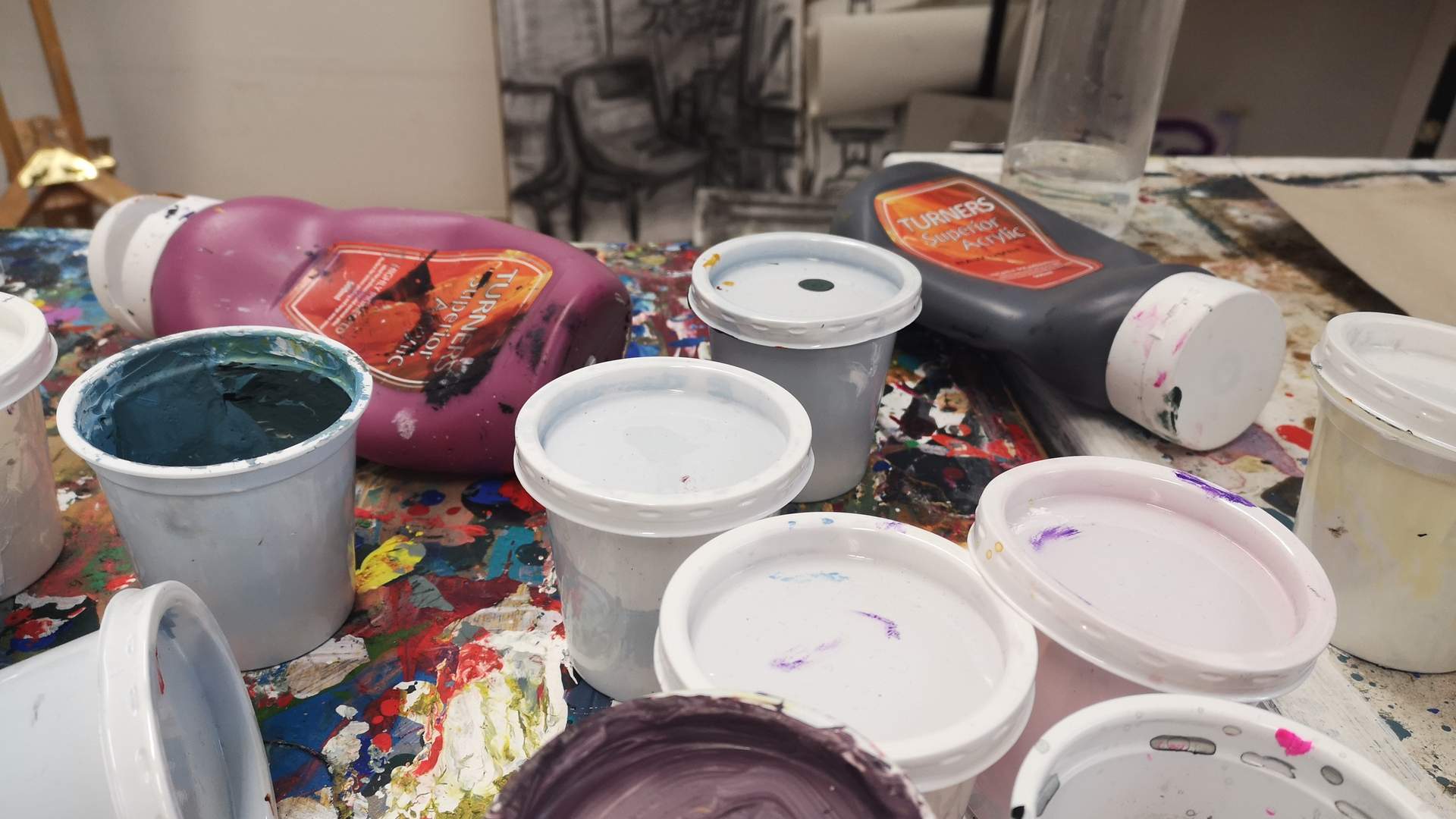A collect of opened and used paint cans sit on a cluttered table filled with art supplies