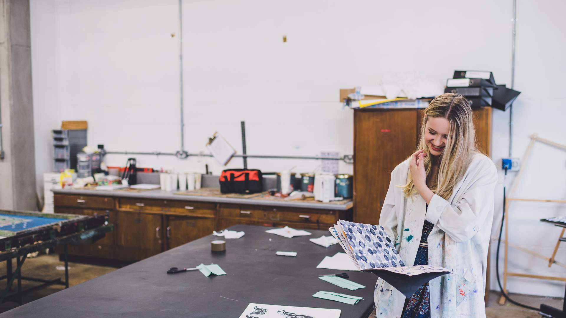 Three women wearing paint-splattered lab coats work together in a classroom. One woman, in a green shirt, smiles and looks down. The other two women, one wearing a hijab, also smile while focusing on what theyre doing. Art supplies are visible.