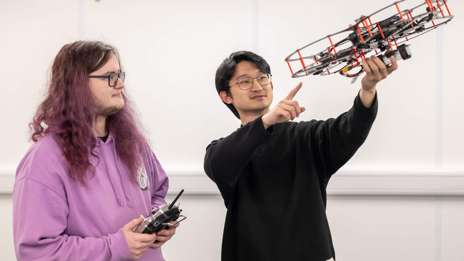 A person with long hair is sitting at a table with a laptop, interacting with a humanoid robot. They appear to be adjusting or programming the robot. The setting is a modern, brightly lit room with yellow chairs.