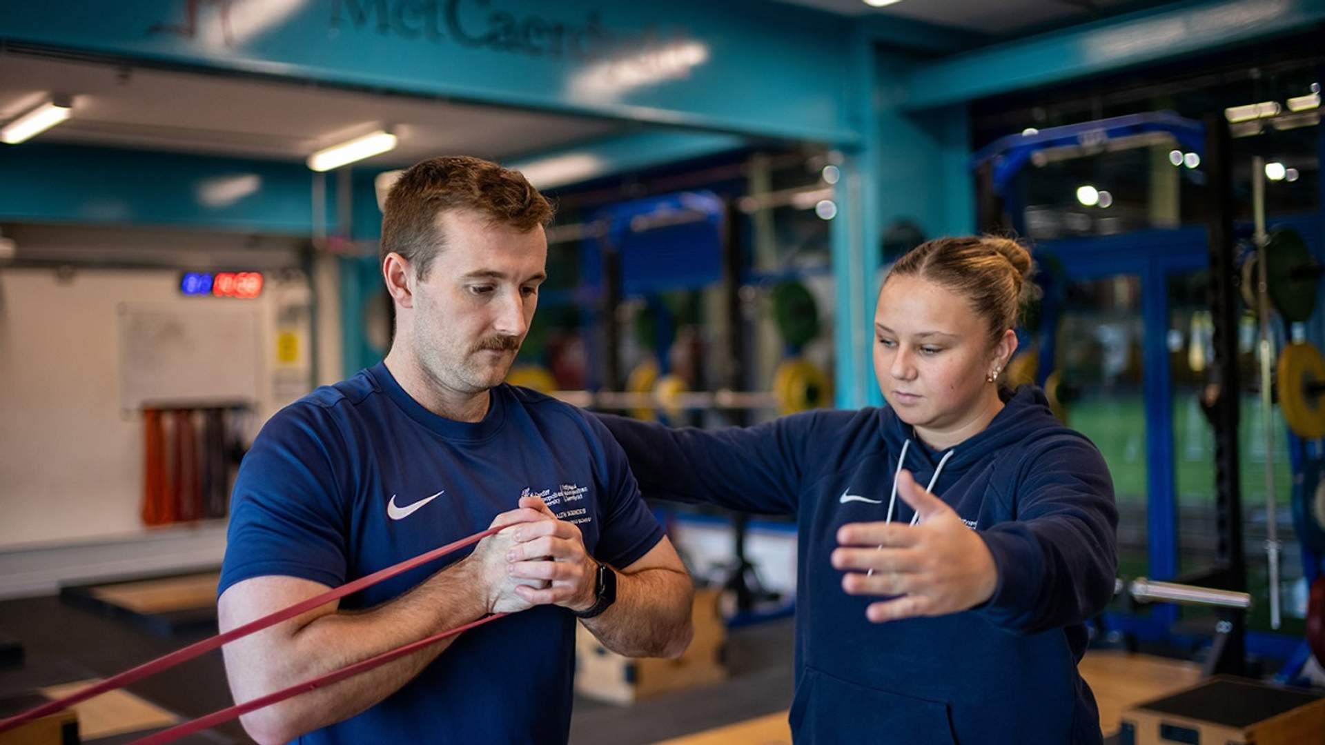 A man and woman exercising in a Cardiff Met Sporty gym, using a resistance band for strength training.