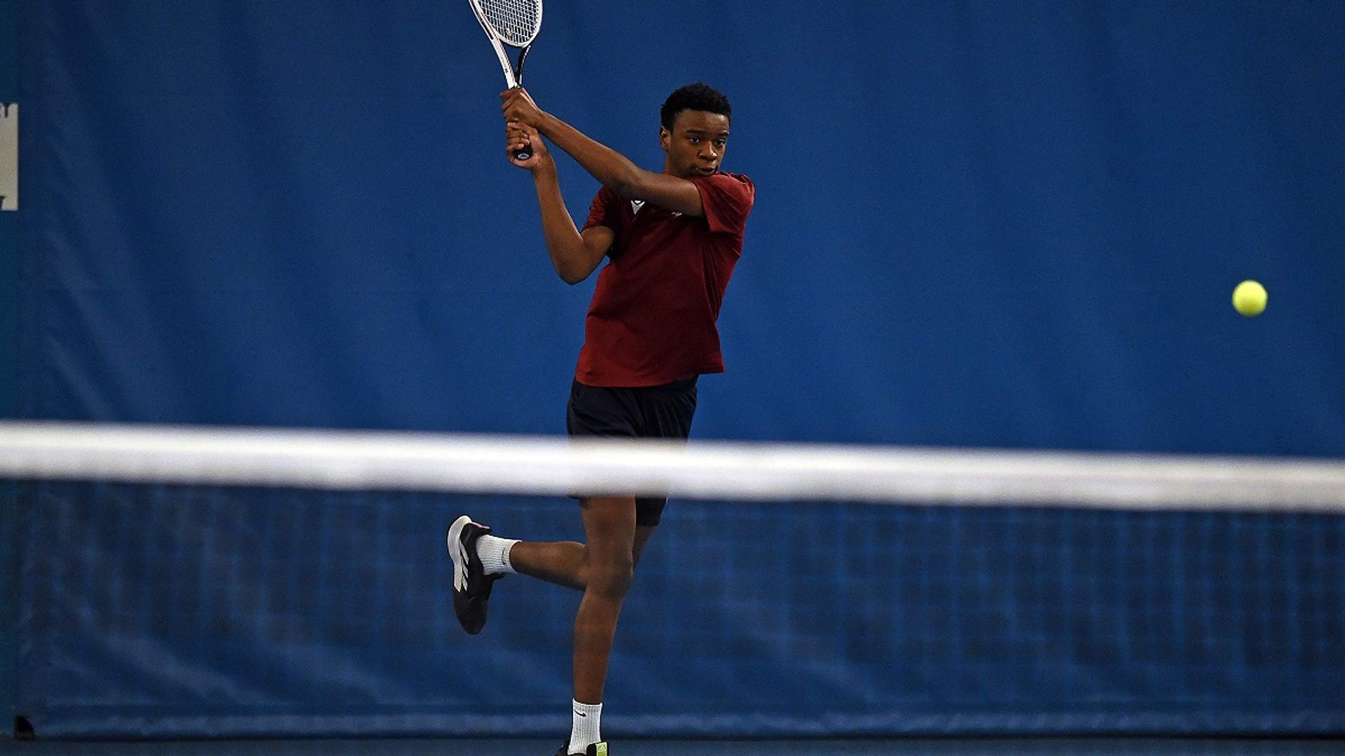 A young man in Cardiff Met exercise t-shirt swings a racket at a tennis ball