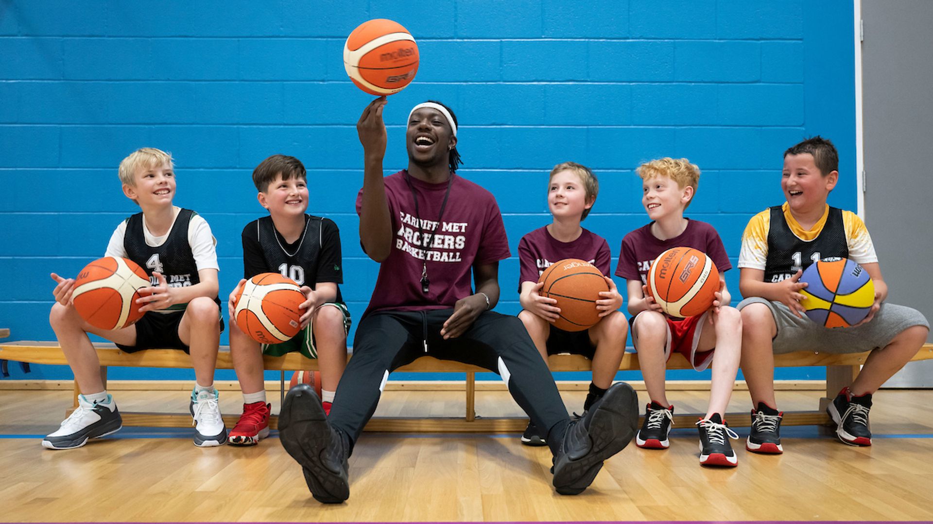 A sports coach spins a basketball on his fingers while four pupils, all holding a basketball on their laps, watch on