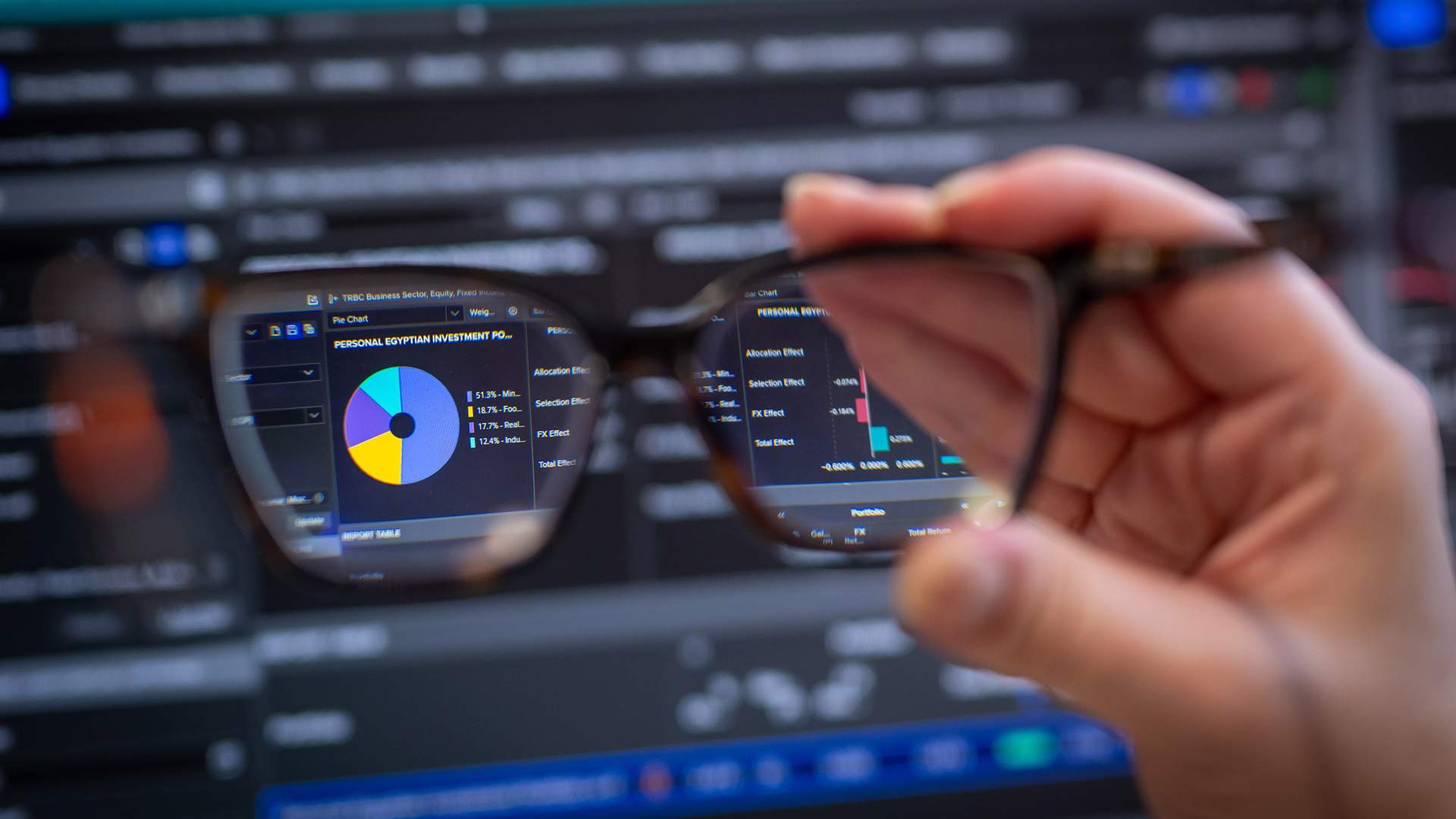 A woman with long blonde hair and glasses is concentrating in front of multiple computer screens displaying financial data. She is wearing a striped shirt and appears focused on her work.