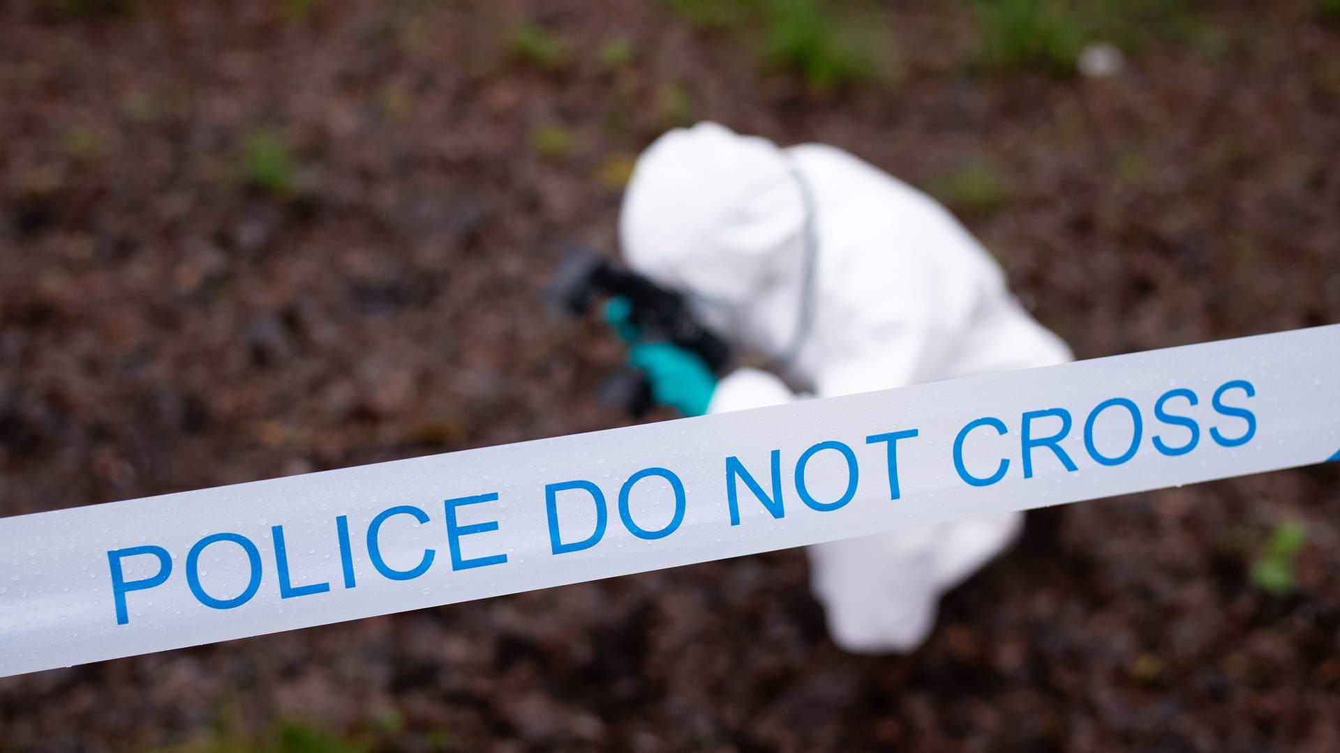 A person with a long ponytail stands outdoors behind blue and white police tape. They are wearing a dark vest with reflective strips and a badge, set against a backdrop of trees and plants.
