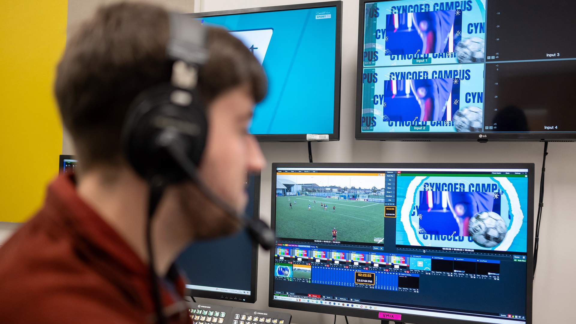 A man in headphones engaged in watching a soccer game on several monitors, showcasing his skills at sports broadcasting.