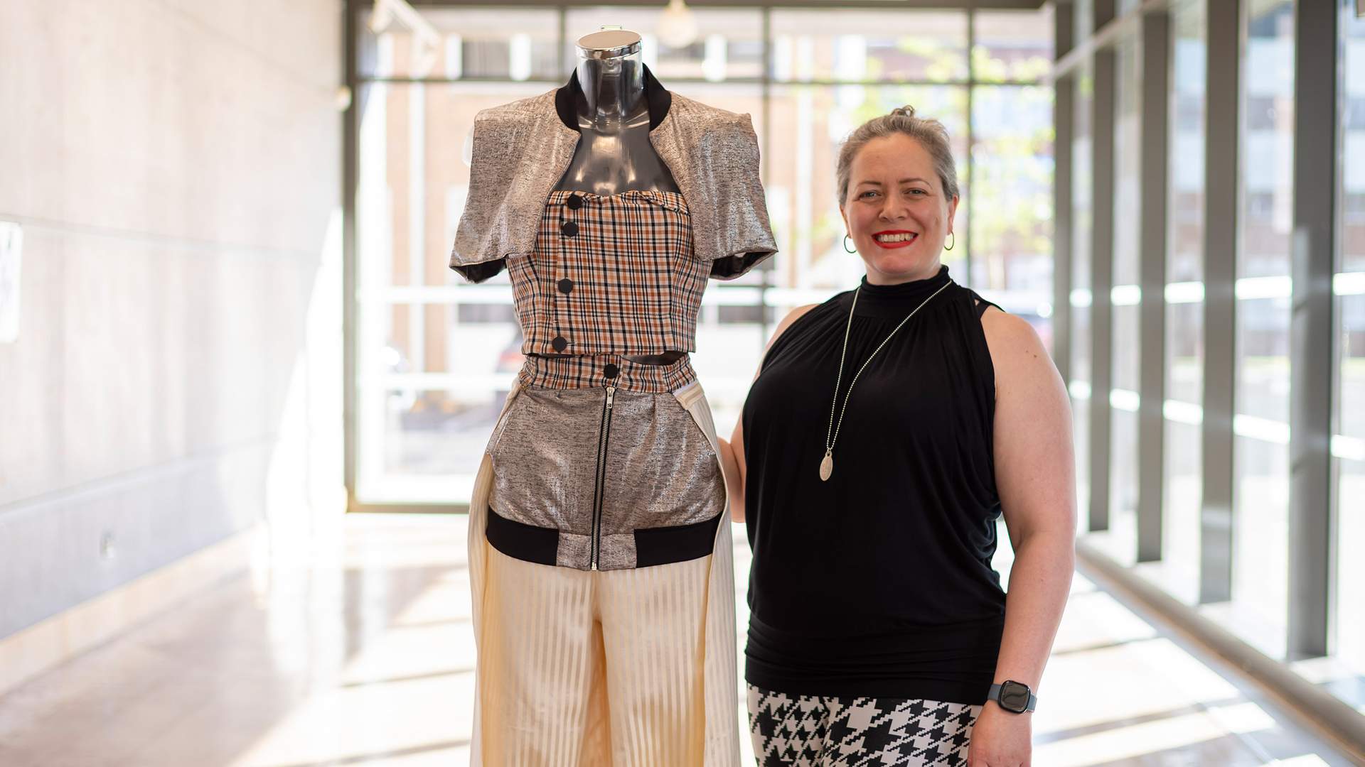A person is focused on working with fabric in a fashion design studio. They are surrounded by dress forms and sewing materials, with others working in the background.