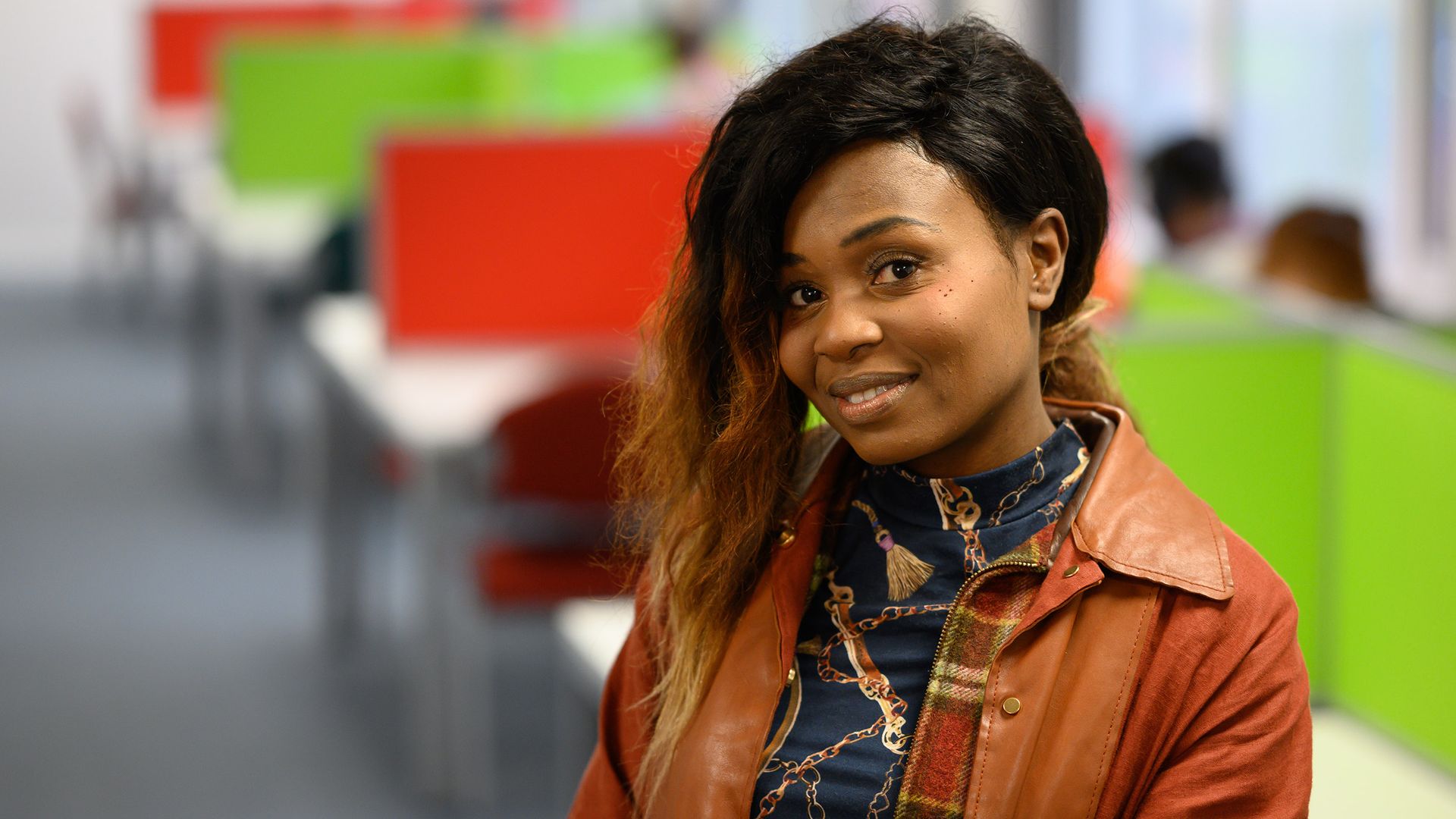 A person smiling and standing with arms crossed in a brightly colored office space. The background features red and green dividers and office furniture.