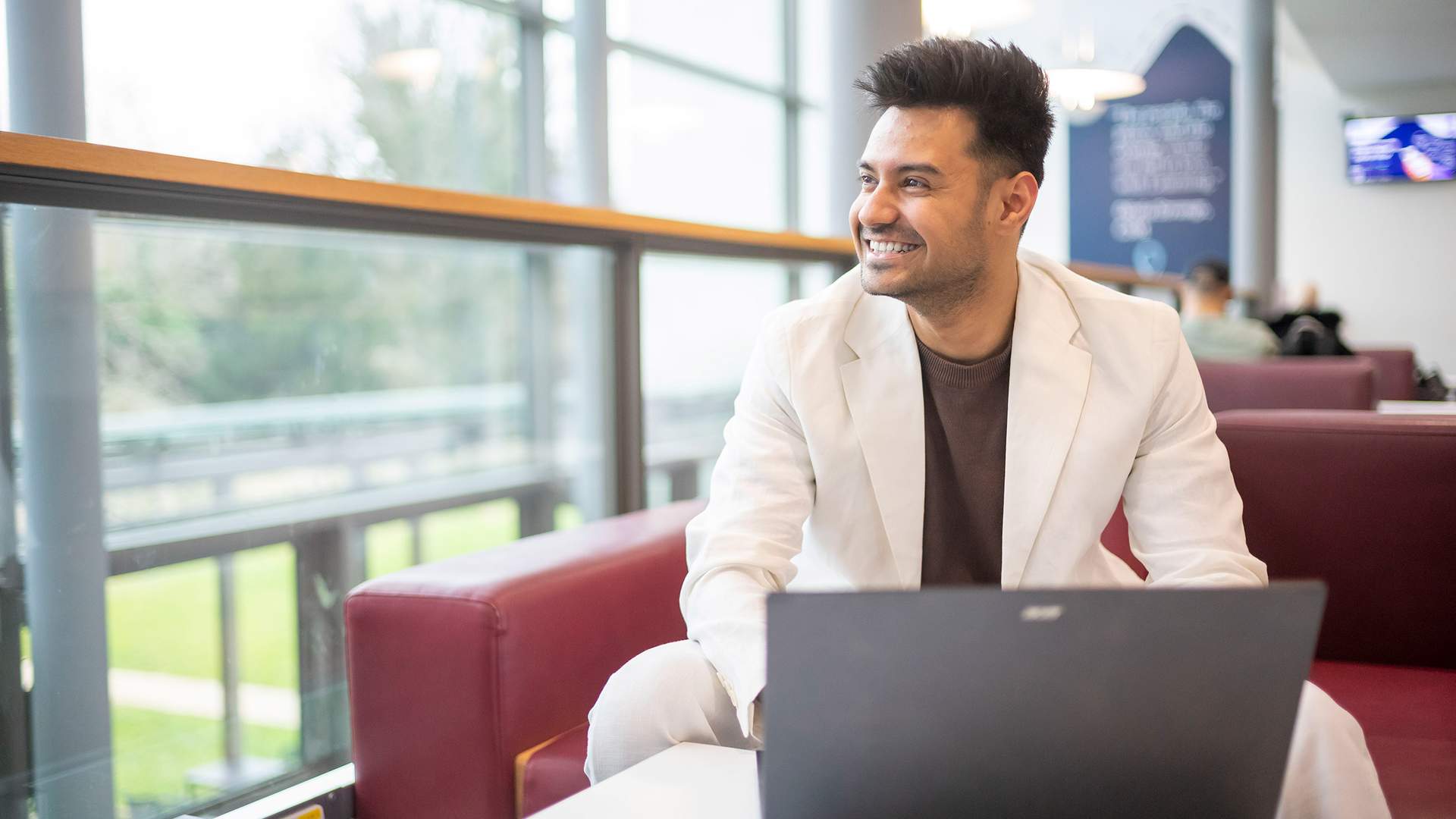 A person with long blonde hair is smiling while seated, wearing a light beige sweater. They are facing another person and have a laptop in front of them on a table. The background has a geometric pattern.