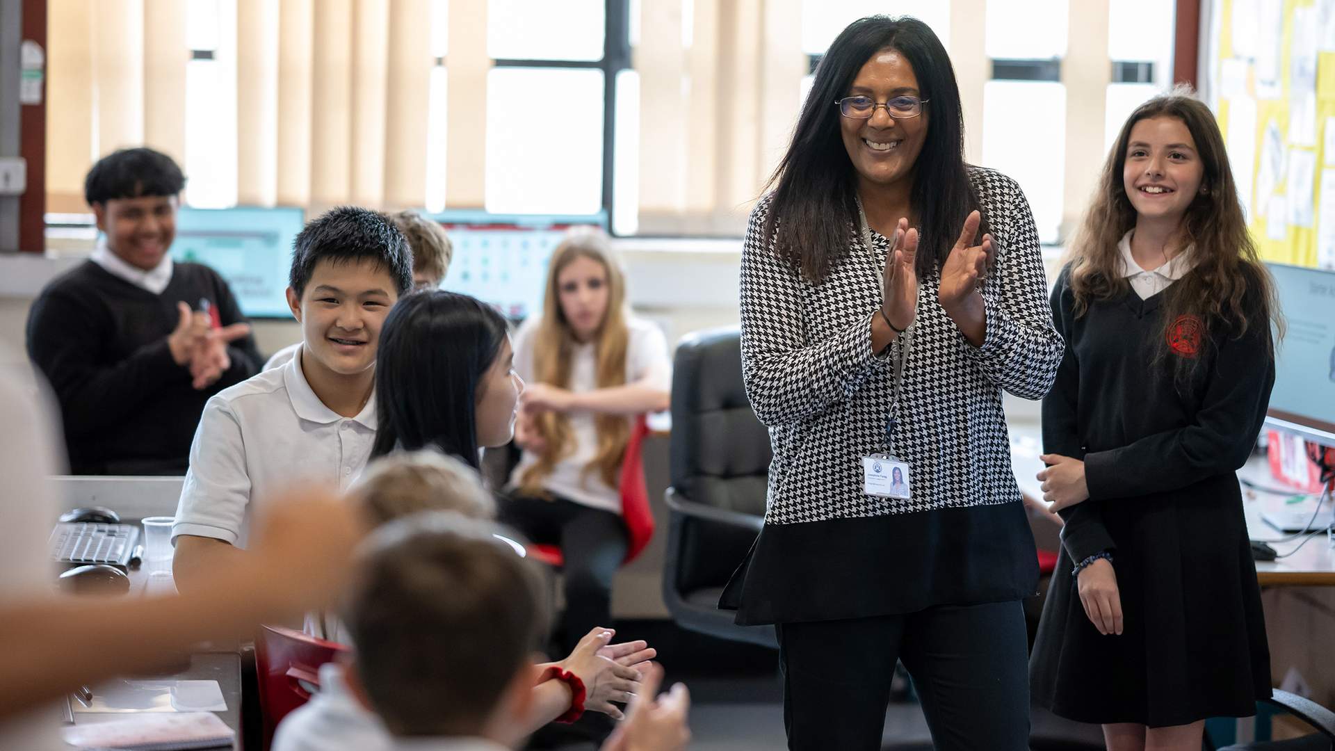 A teacher stands in a classroom surrounded by a group of pupils. There are computer monitors in the background.